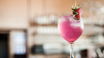ATLANTIC Hotel Airport | Cocktail Pinkes Getränk mit Eis, garniert mit Erdbeere und Limettenscheiben, in einem Glas vor unscharfem Hintergrund.