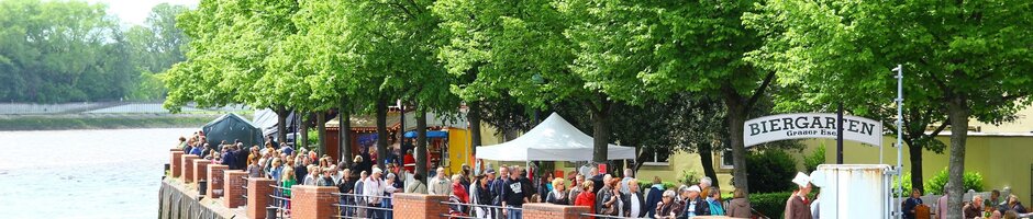 Ansicht auf das Ufer der Weser mit Bäumen während des Vegesacker Hafenfest in Bremen Blick auf belebten Biergarten am Flussufer mit grünen Bäumen und Menschen, die den Ausblick genießen.