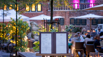 Willkommen im Hofgarten des Restaurant alto Menü auf Staffelei im beleuchteten Gartenrestaurant des ATLANTIC Grand Hotel Bremen, Gäste im Hintergrund.