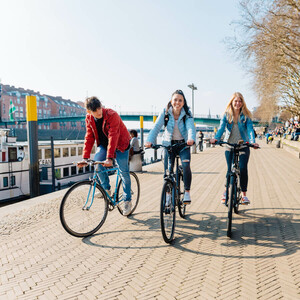 Radwege in Bremen und Umgebung l Bike it Schlachte ©WFB - Jonas Ginter Drei Personen fahren bei sonnigem Wetter auf Fahrrädern entlang einer Uferpromenade, im Hintergrund sind Boote und Gebäude.