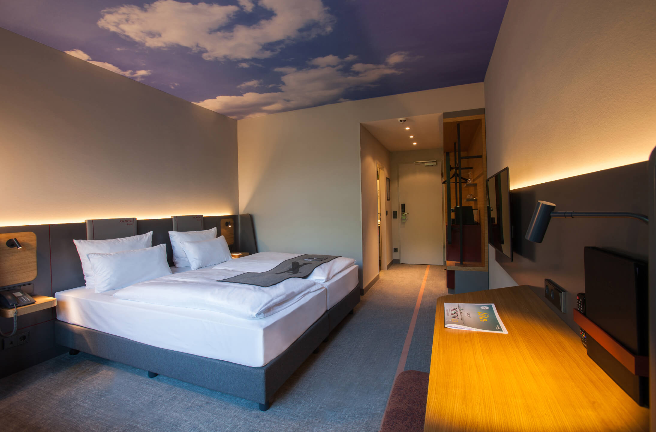 Modernes Hotelzimmer mit Doppelbett, Schreibtisch und Himmelmotiv an der Decke im ATLANTIC Hotel Airport Bremen.