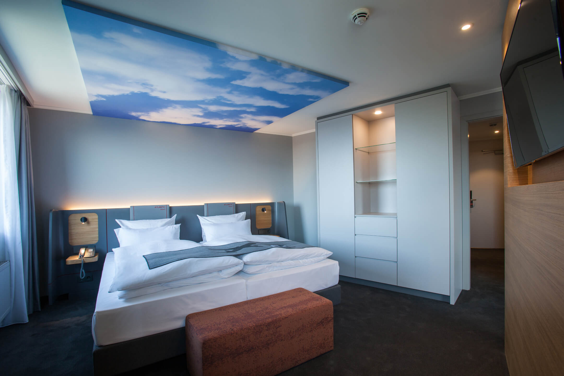 Modernes Hotelzimmer mit Doppelbett, Himmelmotiv an der Decke, Schrank und Sitzbank im ATLANTIC Hotel Airport Bremen.