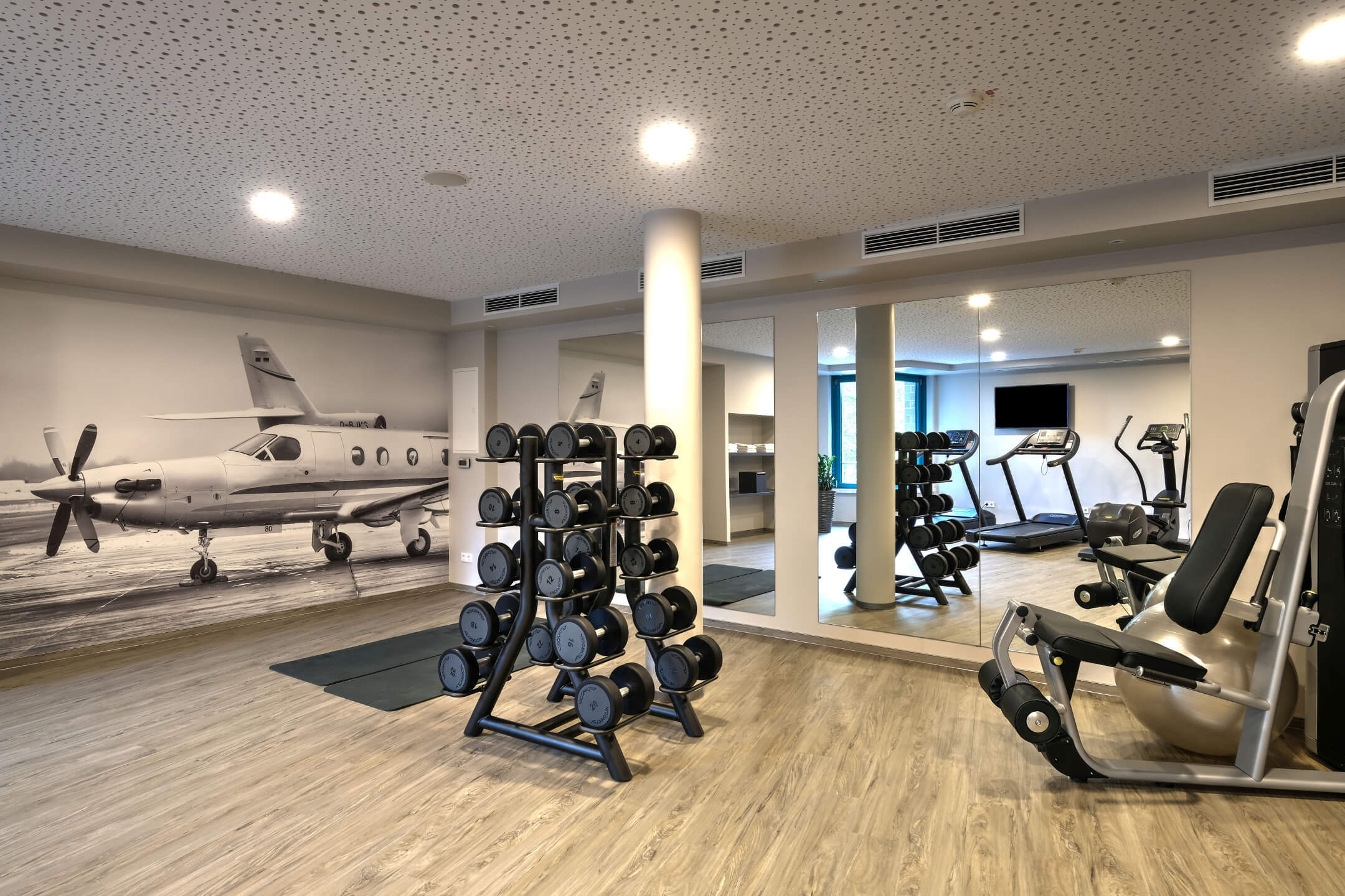Fitnessraum im ATLANTIC Hotel Airport Bremen mit Hanteln, Fitnessgeräten und Flugzeugbild an der Wand.