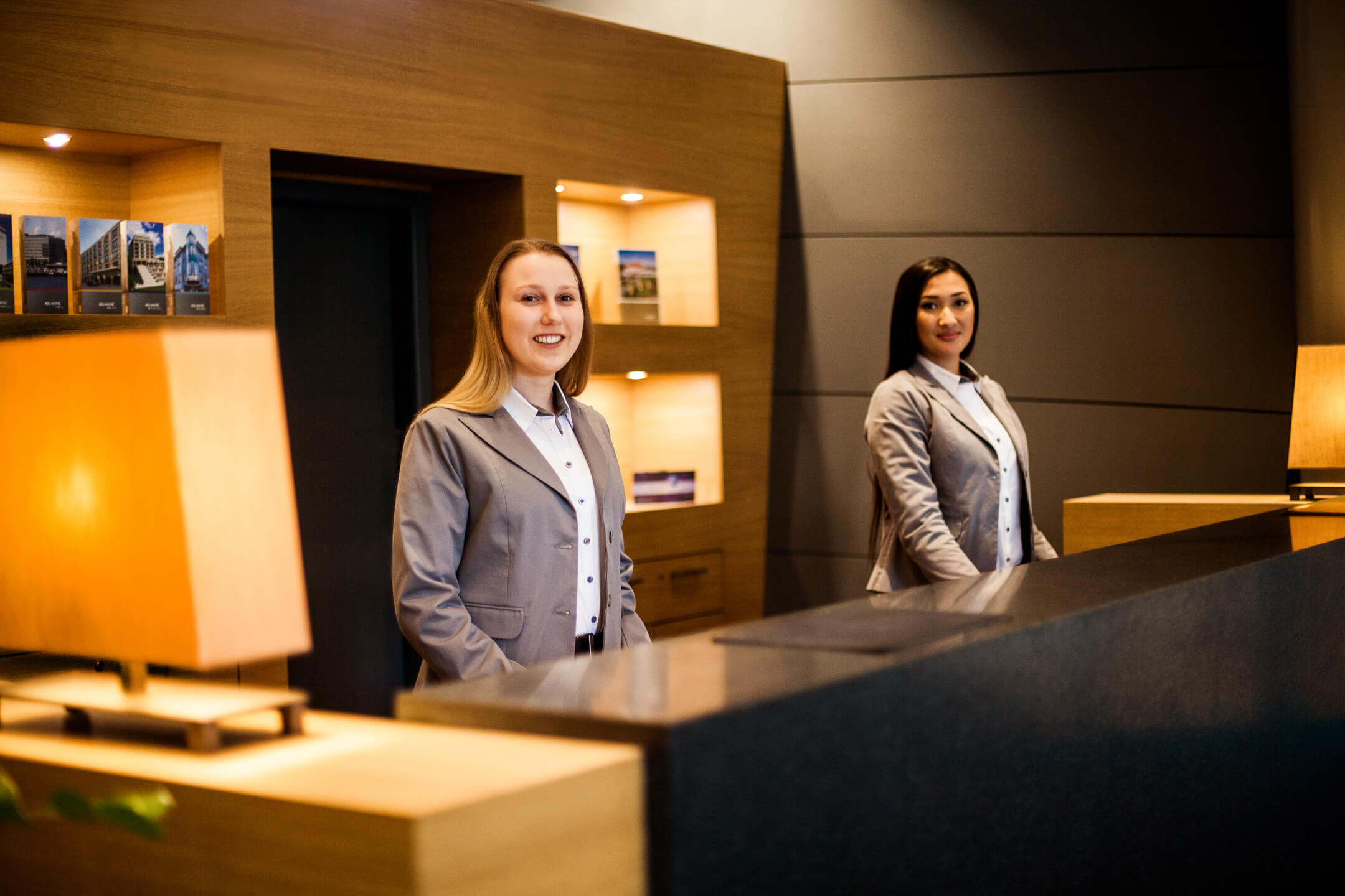 Zwei Empfangsmitarbeiterinnen im ATLANTIC Hotel Airport Bremen lächeln an der Rezeption freundlich.