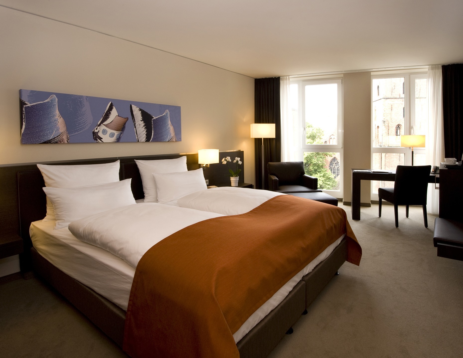 Modern hotel room with double bed, orange ceiling, desk, armchairs and large window with a view of the church.