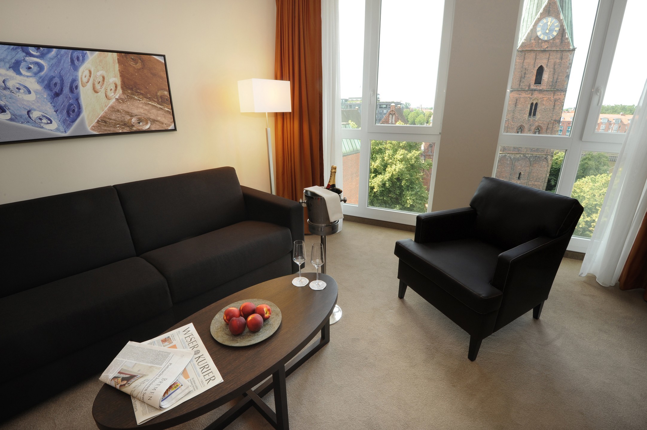 Modern hotel room with sofa, armchair, coffee table, champagne bottle and view of Bremen's church tower through large windows.