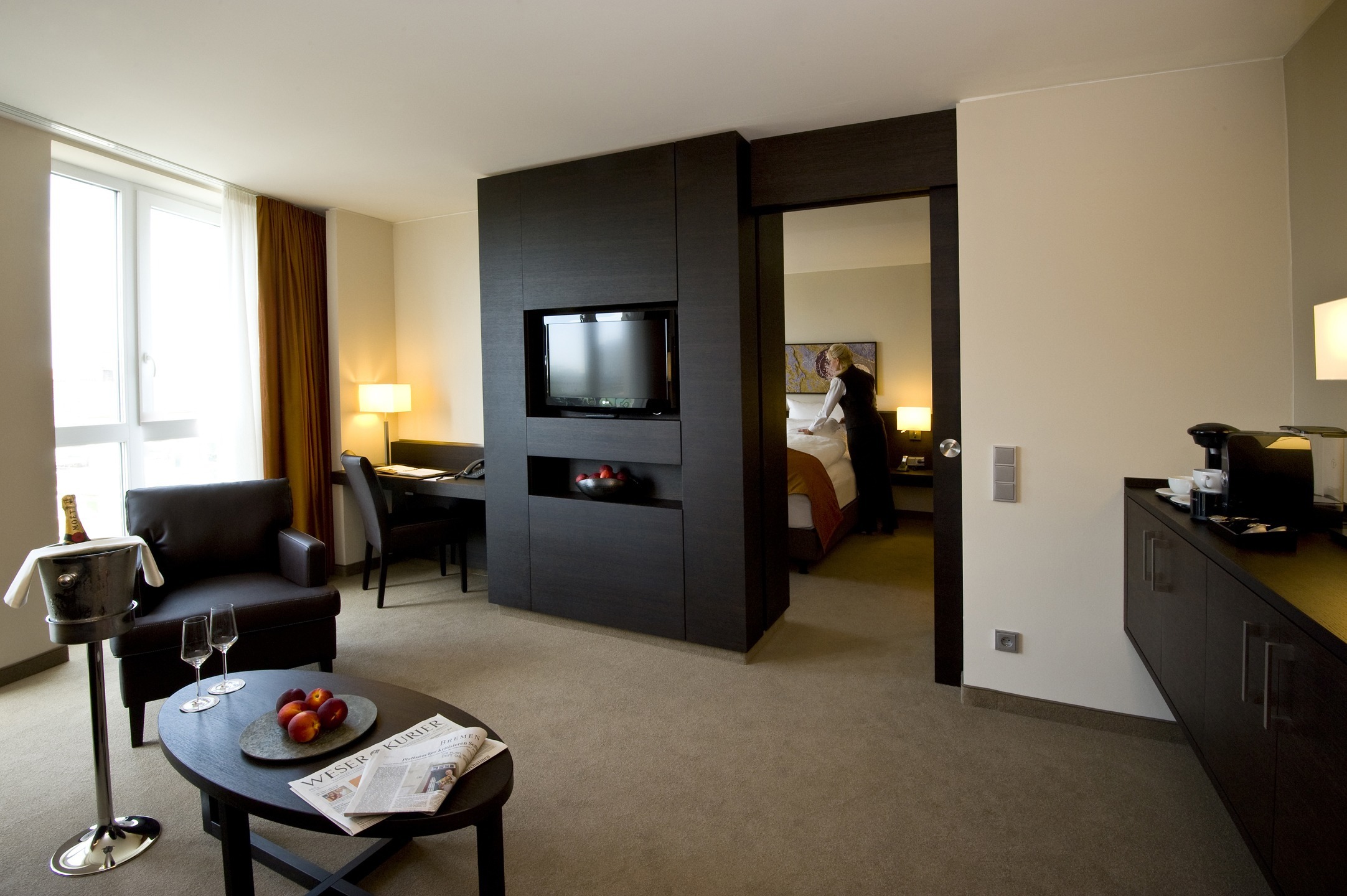 Modern hotel room with seating area, desk, flat-screen TV and view into the bedroom.
