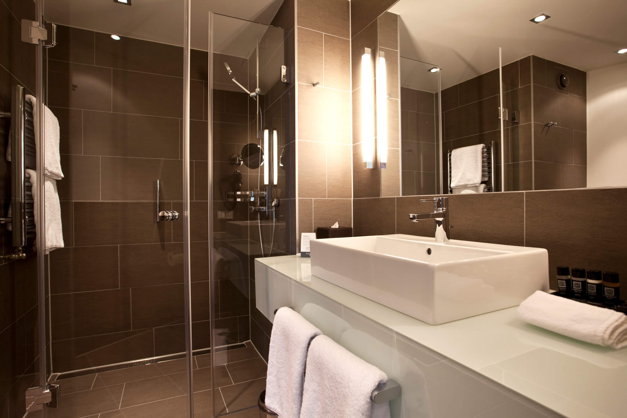 Modern bathroom in the ATLANTIC Grand Hotel Bremen with glass shower, washbasin and towels on a light shelf.
