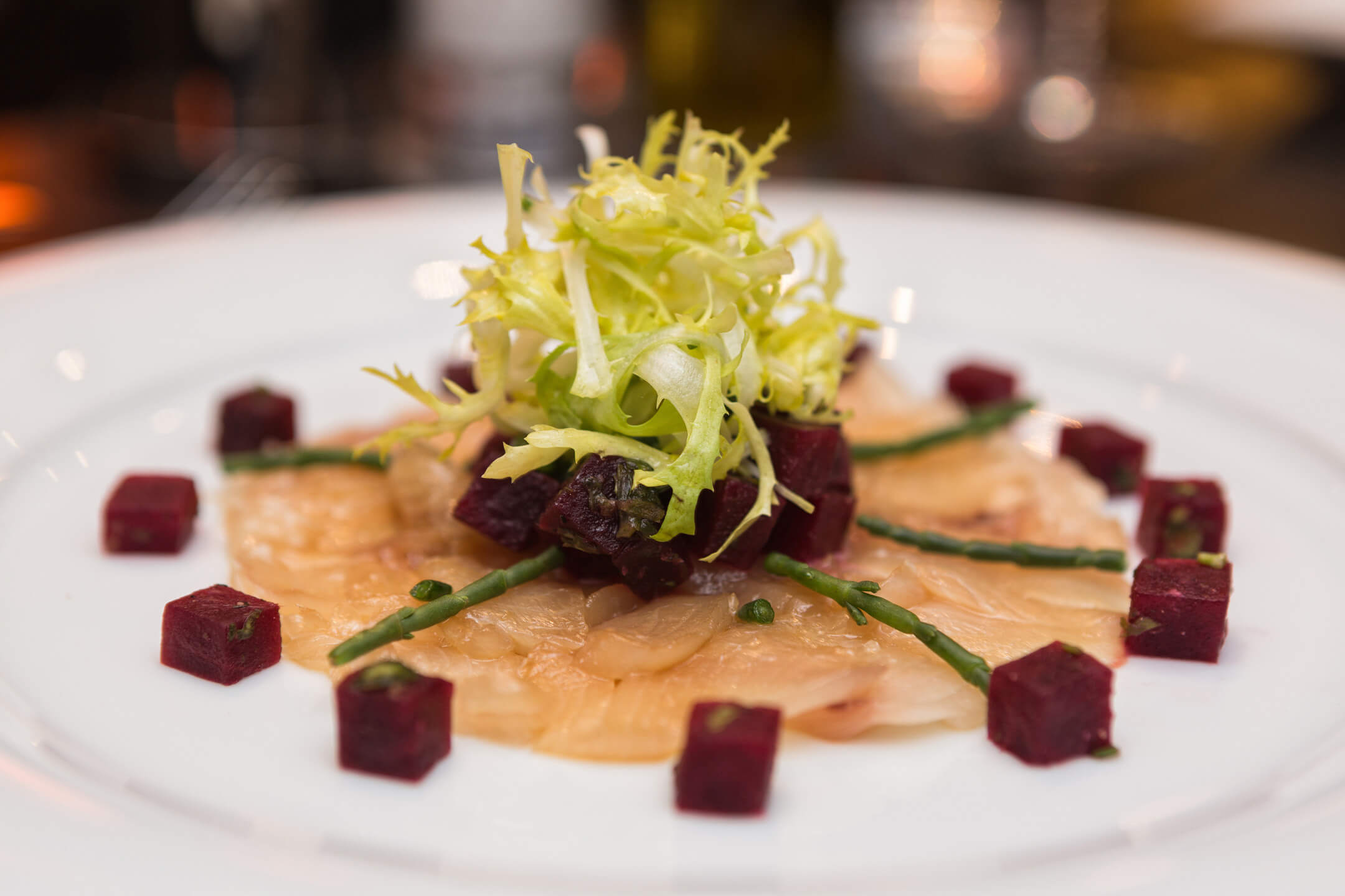Gourmet starter with salmon carpaccio, beet cubes, salicorns and frisée salad on a white plate.