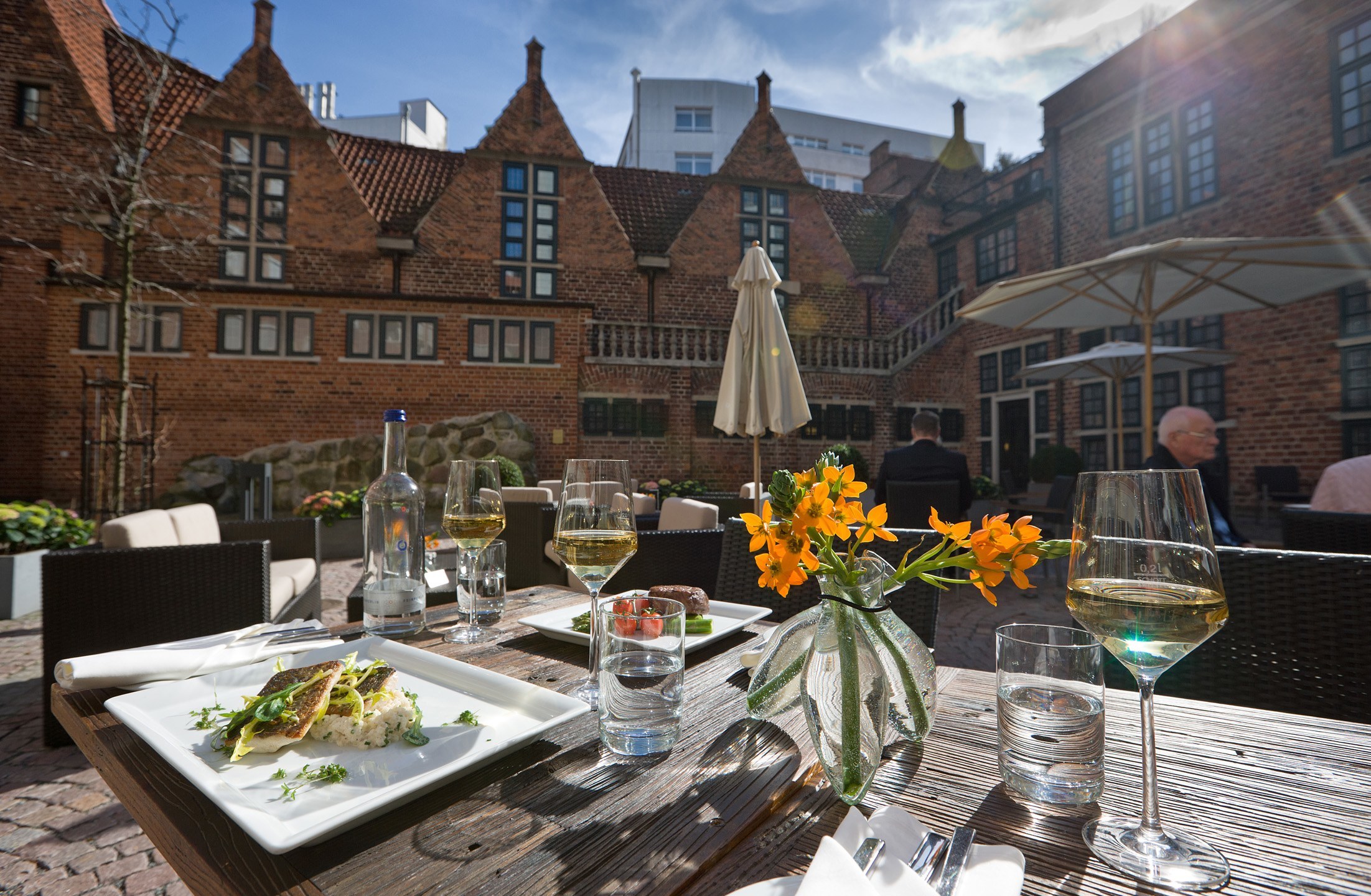 Well-kept hotel courtyard with laid table, wine, flowers, historic brick façade and parasols in the background.