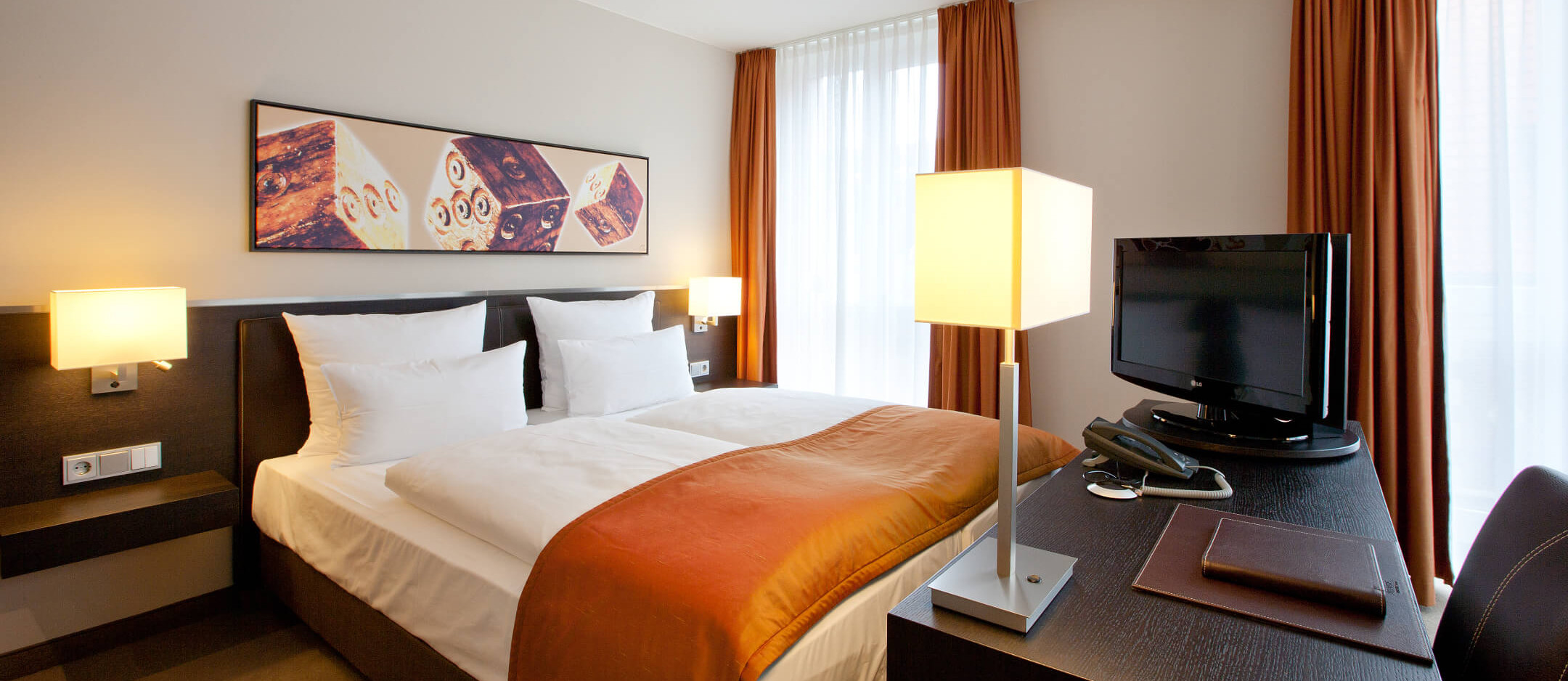 Modernes Hotelzimmer mit Doppelbett, orangefarbener Tagesdecke, Schreibtisch, Flachbild-TV und stilvoller Beleuchtung.