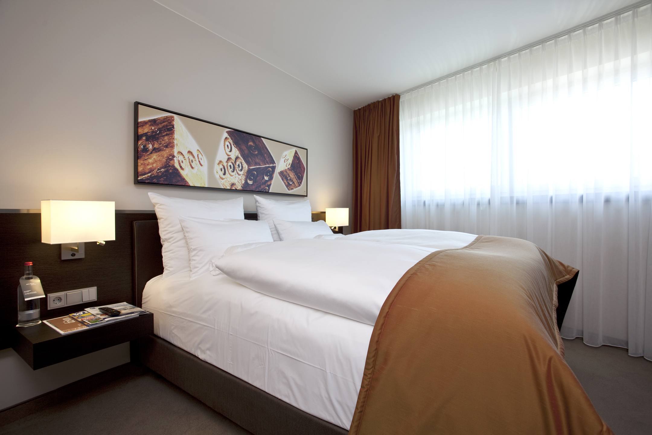 Modernes Hotelzimmer mit Doppelbett, weißer Bettwäsche, brauner Tagesdecke, Kunstwerk an der Wand und Tageslicht durch Fenster.