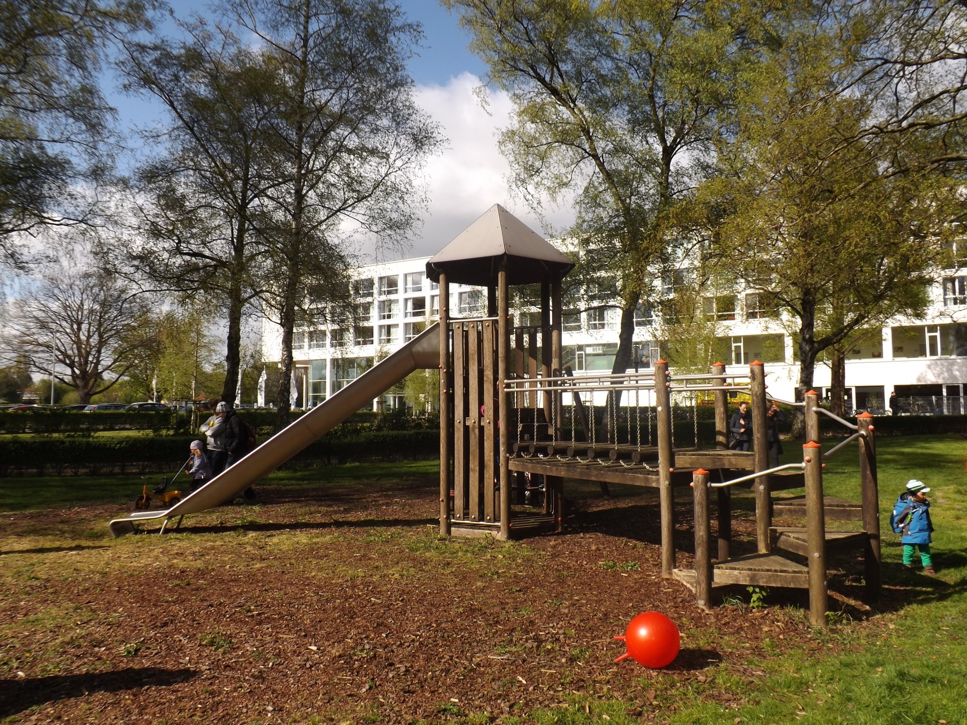 Playground with slide and climbing frame in front of a hotel building, surrounded by trees.