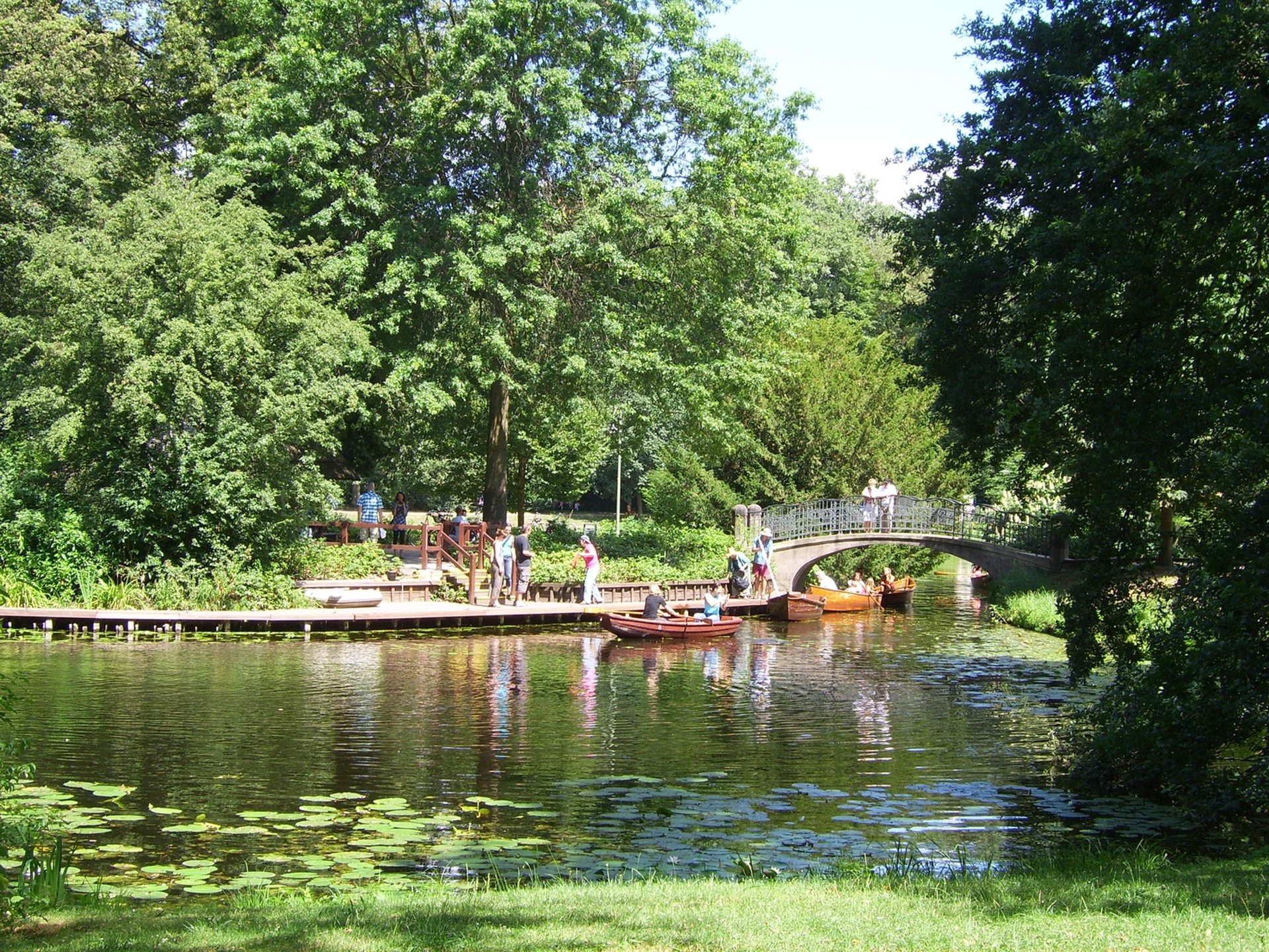 Ruhiger Fluss mit Holzbooten, umgeben von üppigem Grün, Fußgängerbrücke im Hintergrund.