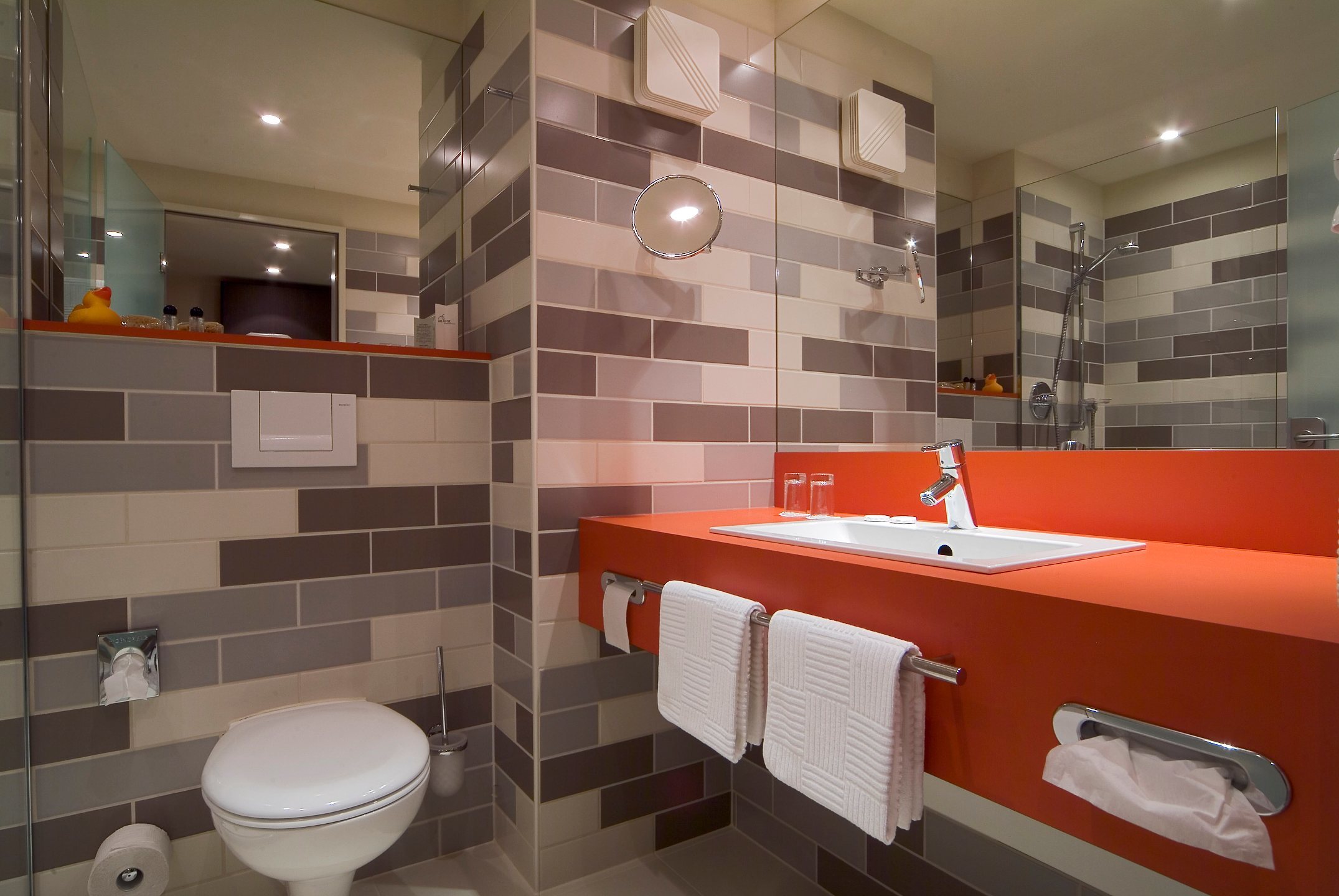 Modernes Badezimmer mit orangefarbenem Waschtisch, Spiegel, Handtüchern und Dusche im ATLANTIC Hotel Galopprennbahn Bremen.