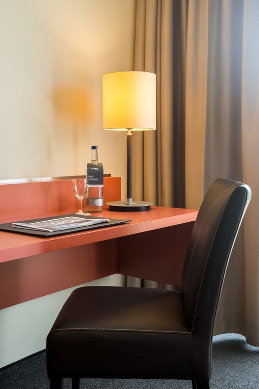 Schreibtisch mit Lampe, Wasserflasche und Glas im ATLANTIC Hotelzimmer, brauner Stuhl und Vorhänge im Hintergrund.