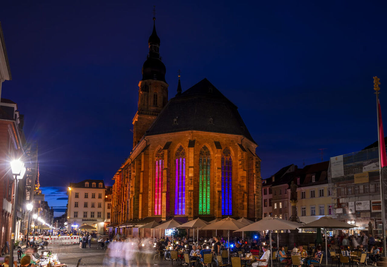 Lively square at night, church with colorful lighting, people sitting under umbrellas, cozy atmosphere.