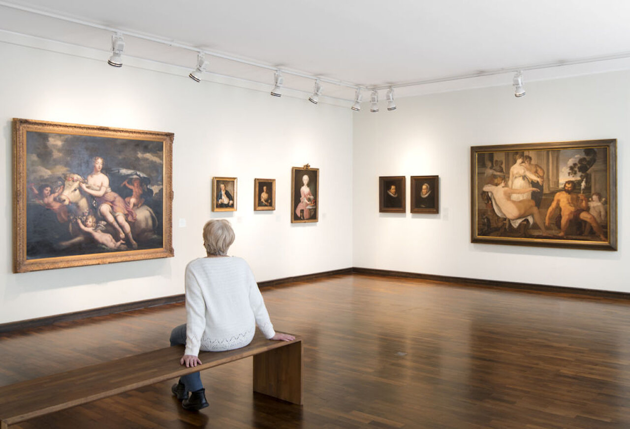 Person sitting on a bench in an art gallery, surrounded by classical paintings on white walls.