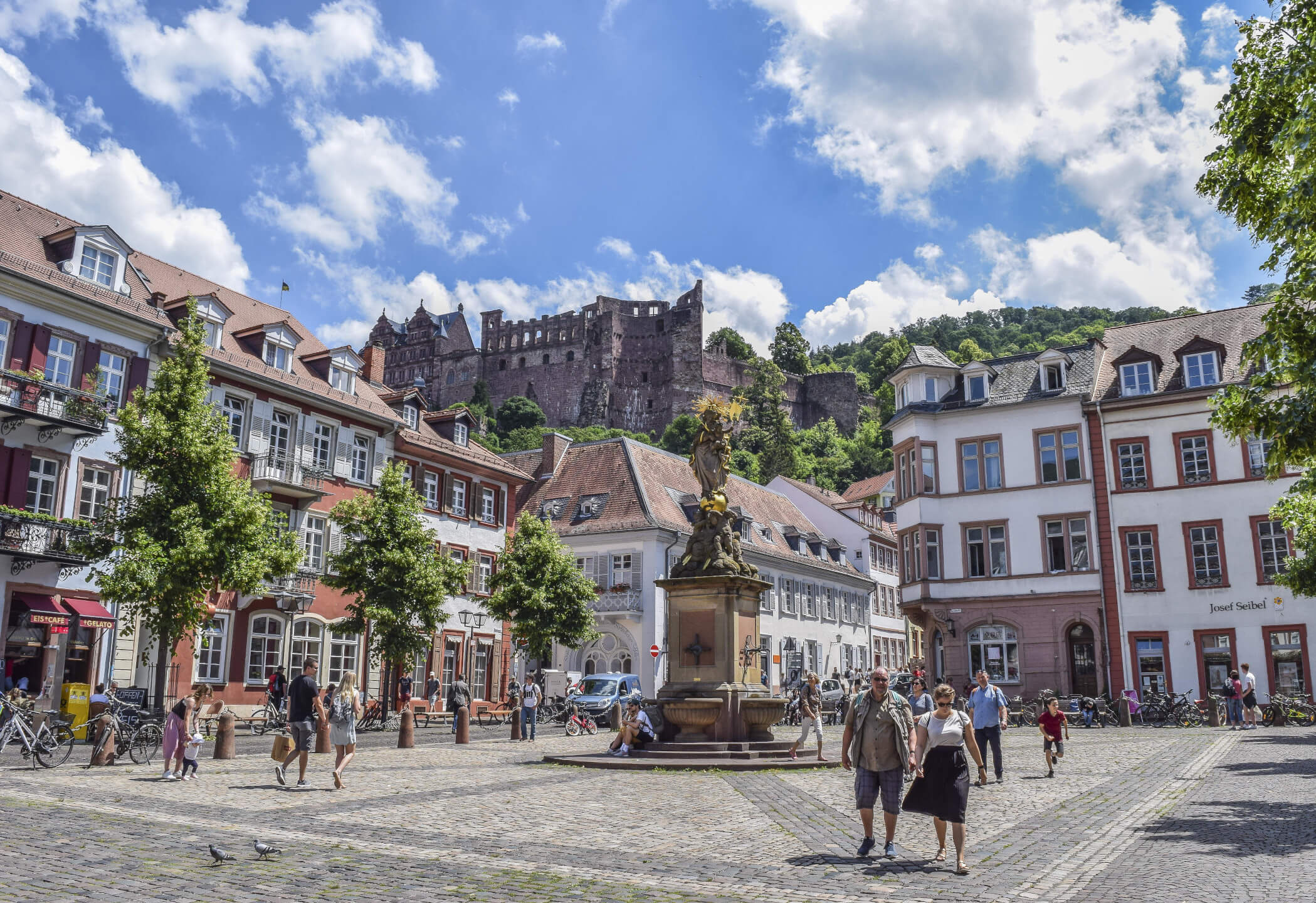 Historic square with fountain, surrounded by old buildings and trees, with a castle on a hill in the background.