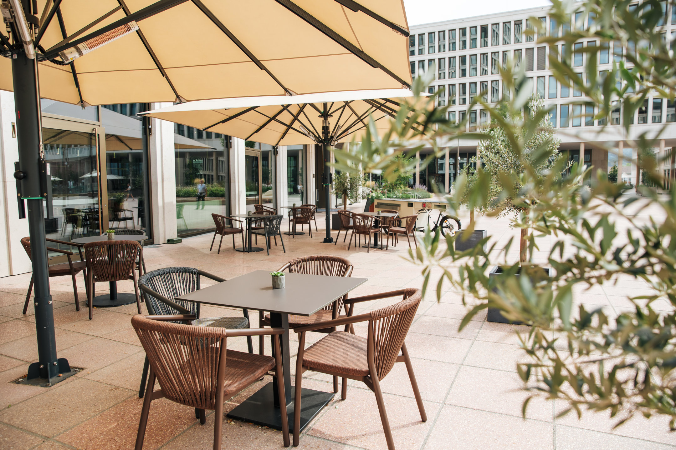 Die Terrasse der Deli Lounge mit beigen Sonnenschirmen und mehreren Tischen