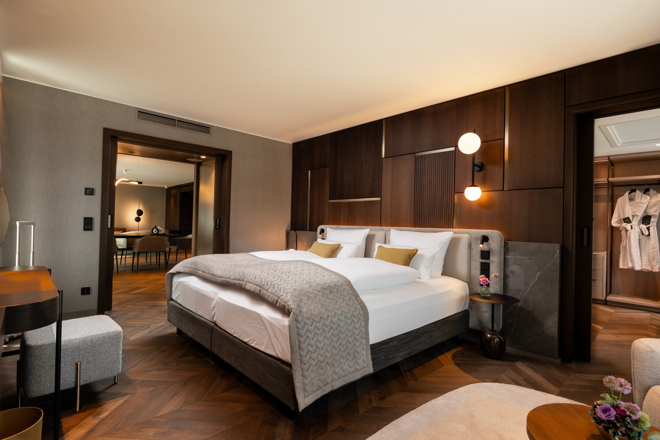 Modernes Hotelzimmer mit Doppelbett, Holzwänden, und Blick in einen Wohnbereich. Elegantes Design mit warmen Farben.