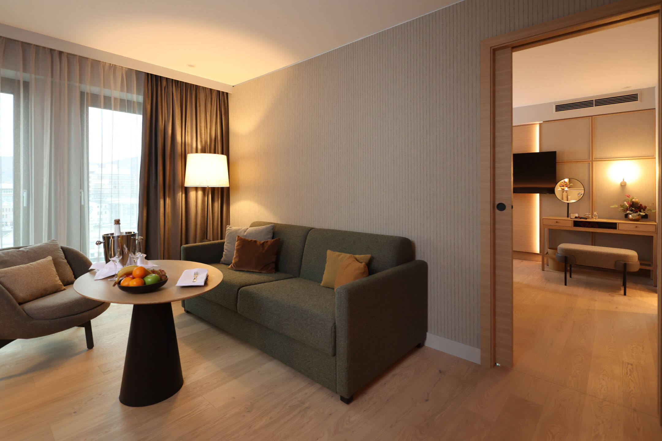Modern hotel room with cozy sitting area, sofa, table with fruit, lamp and view of the adjoining room with TV.