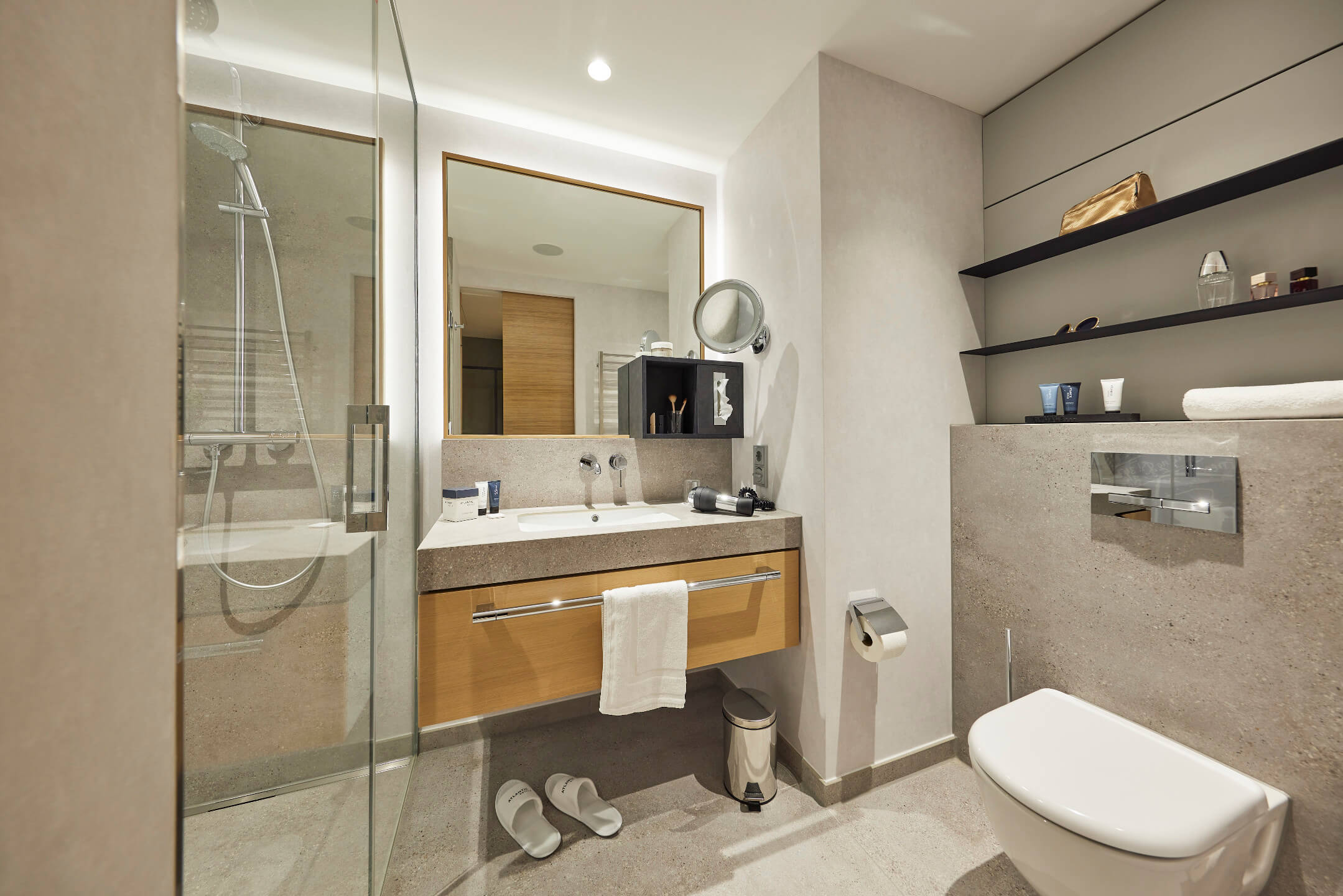 Modernes Hotelbadezimmer mit Dusche, Waschbecken, Spiegel, WC und Regalen mit Pflegeprodukten im ATLANTIC Hotel Heidelberg.