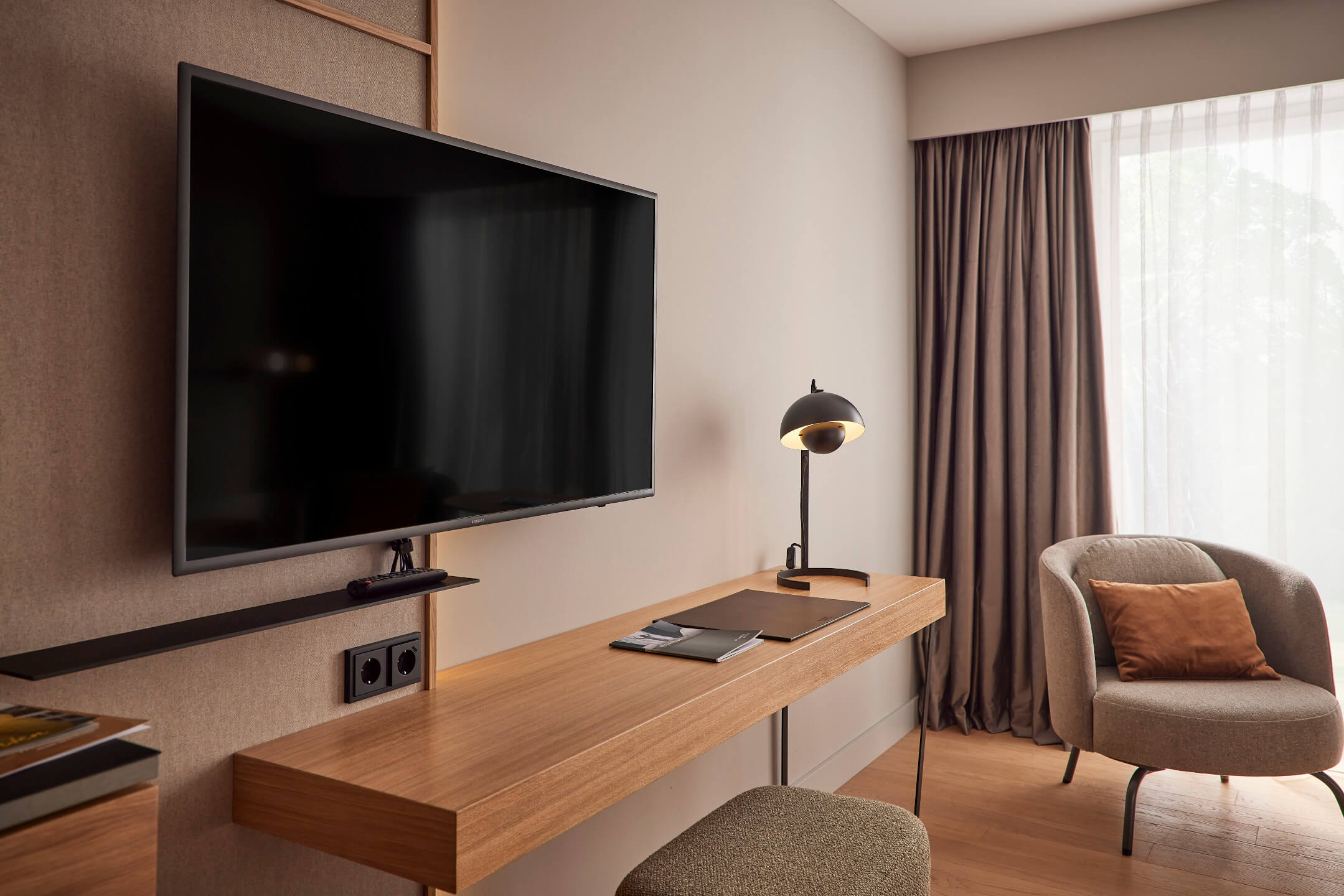 Modernes Hotelzimmer mit Schreibtisch, Lampe, Fernseher und gemütlichem Sessel vor einem Fenster mit Vorhängen.