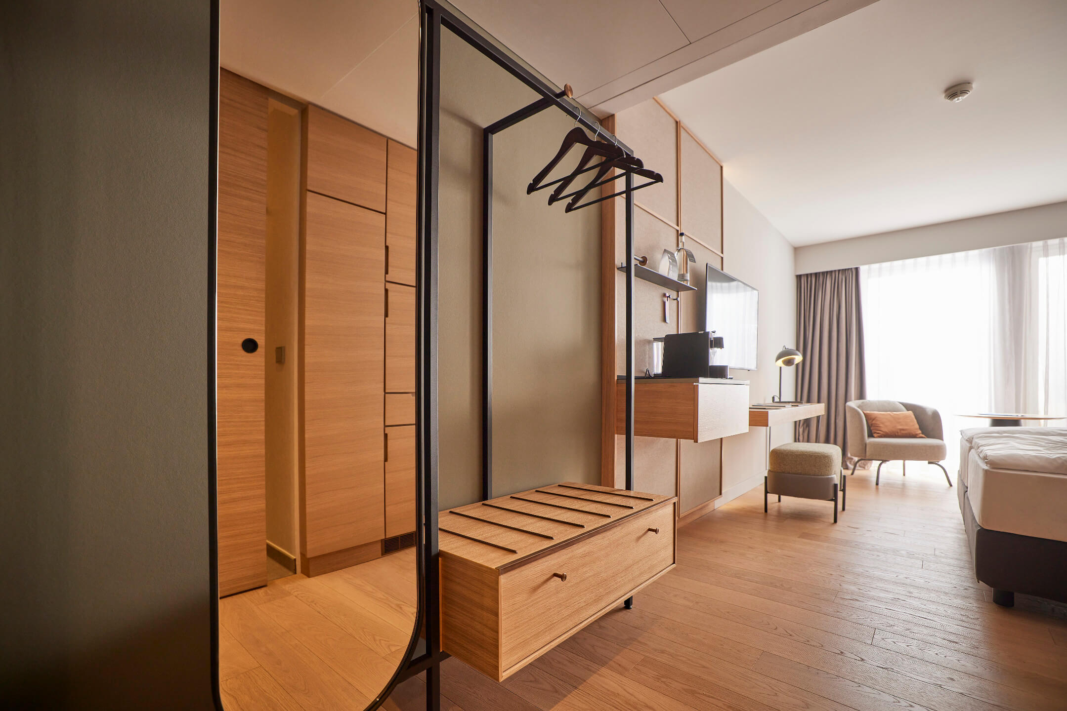 Modernes Hotelzimmer mit Holzfußboden, Doppelbett, Schreibtisch, Sessel und großem Fenster mit Vorhängen.