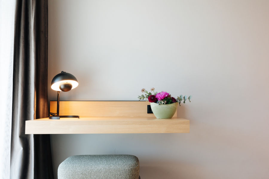 Minimalist hotel room with wooden table, table lamp and flower vase, next to a gray stool.