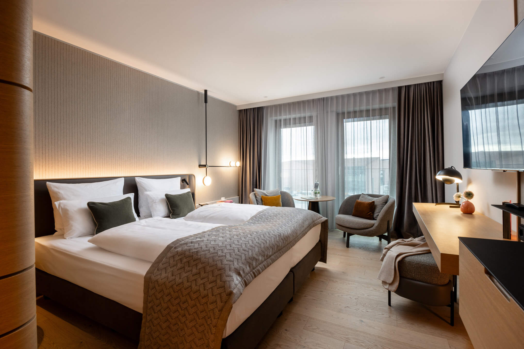 Modernes Hotelzimmer mit Doppelbett, Schreibtisch, zwei Sesseln und großen Fenstern mit Vorhängen im ATLANTIC Hotel Heidelberg.