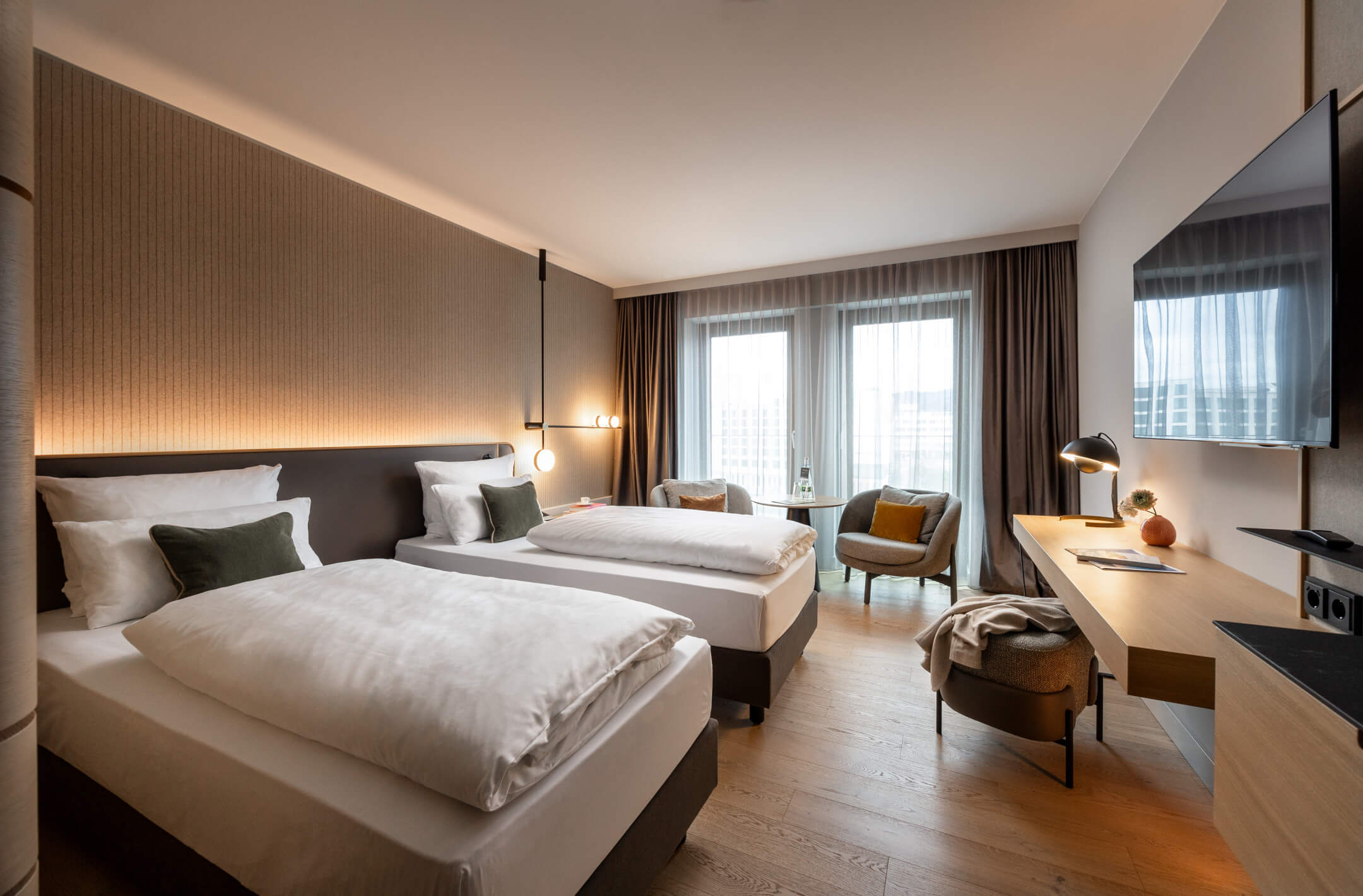 Modernes Hotelzimmer mit zwei Einzelbetten, Schreibtisch, Sessel und großen Fenstern mit Vorhängen im ATLANTIC Hotel Heidelberg.