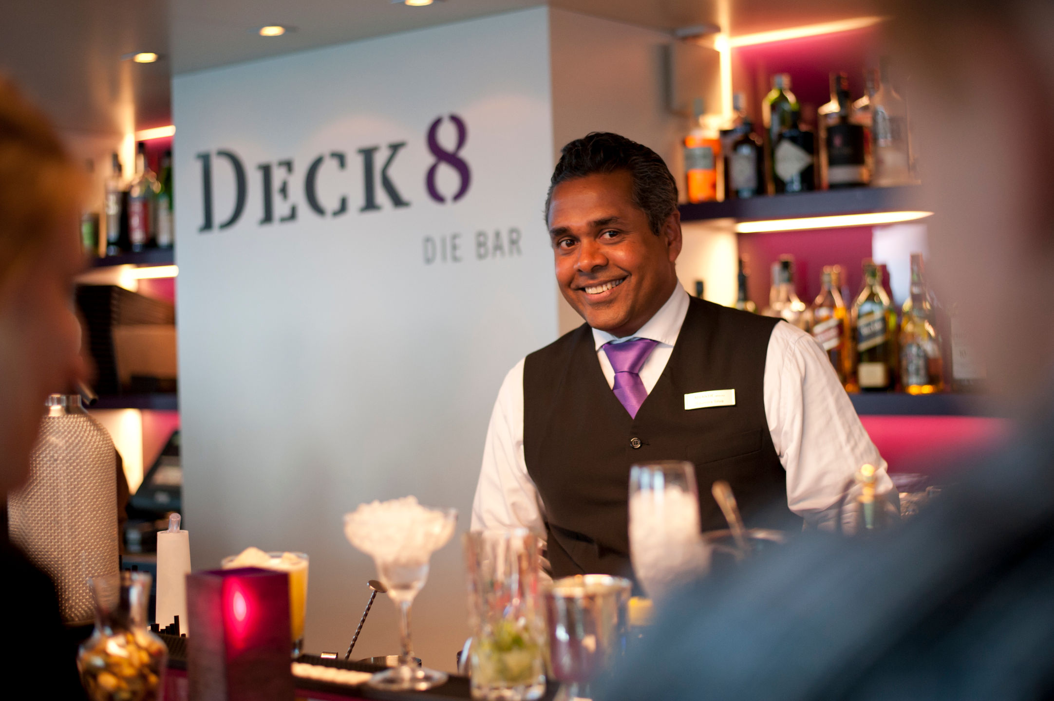 ATLANTIC Hotel Kiel – Bar tender Bartender in an elegant vest smiles behind the counter of the stylish "Deck 8" bar, surrounded by bottles and glasses.