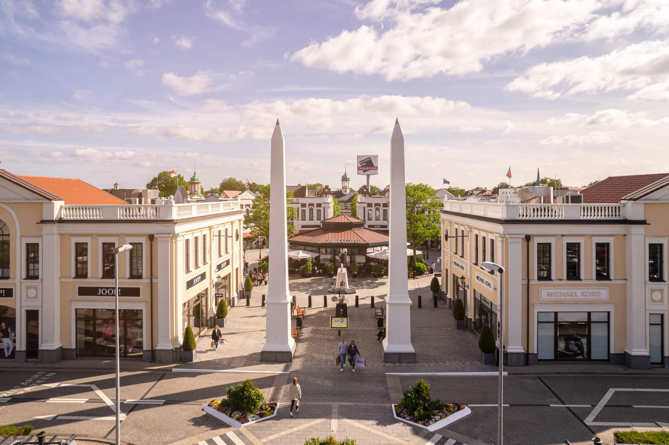 Designer Outlet Neumünster Shopping street with two white obelisks, surrounded by elegant buildings and stores under a blue sky.