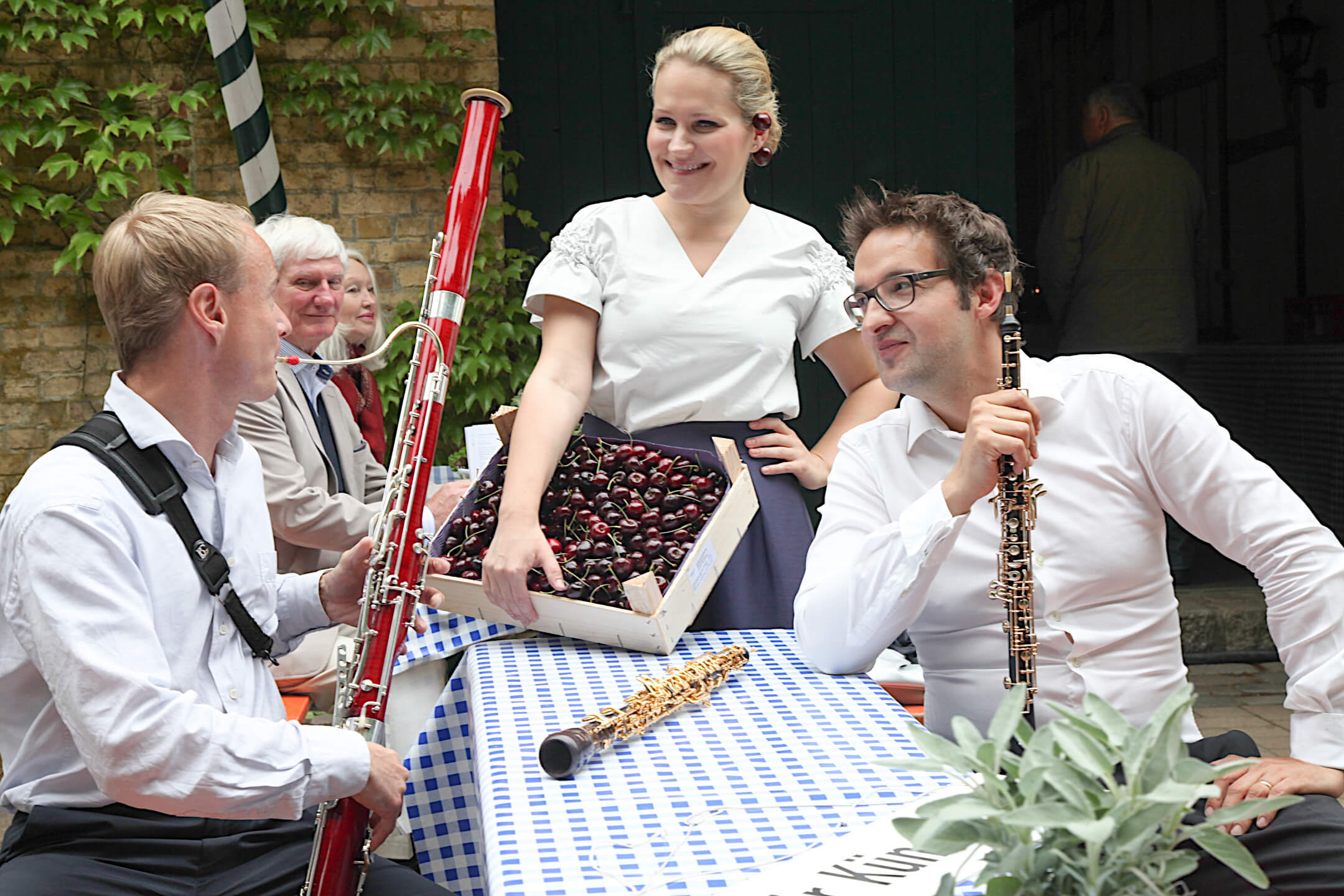 People at a party, two men with instruments and a woman with cherries, at a table with a blue and white tablecloth.