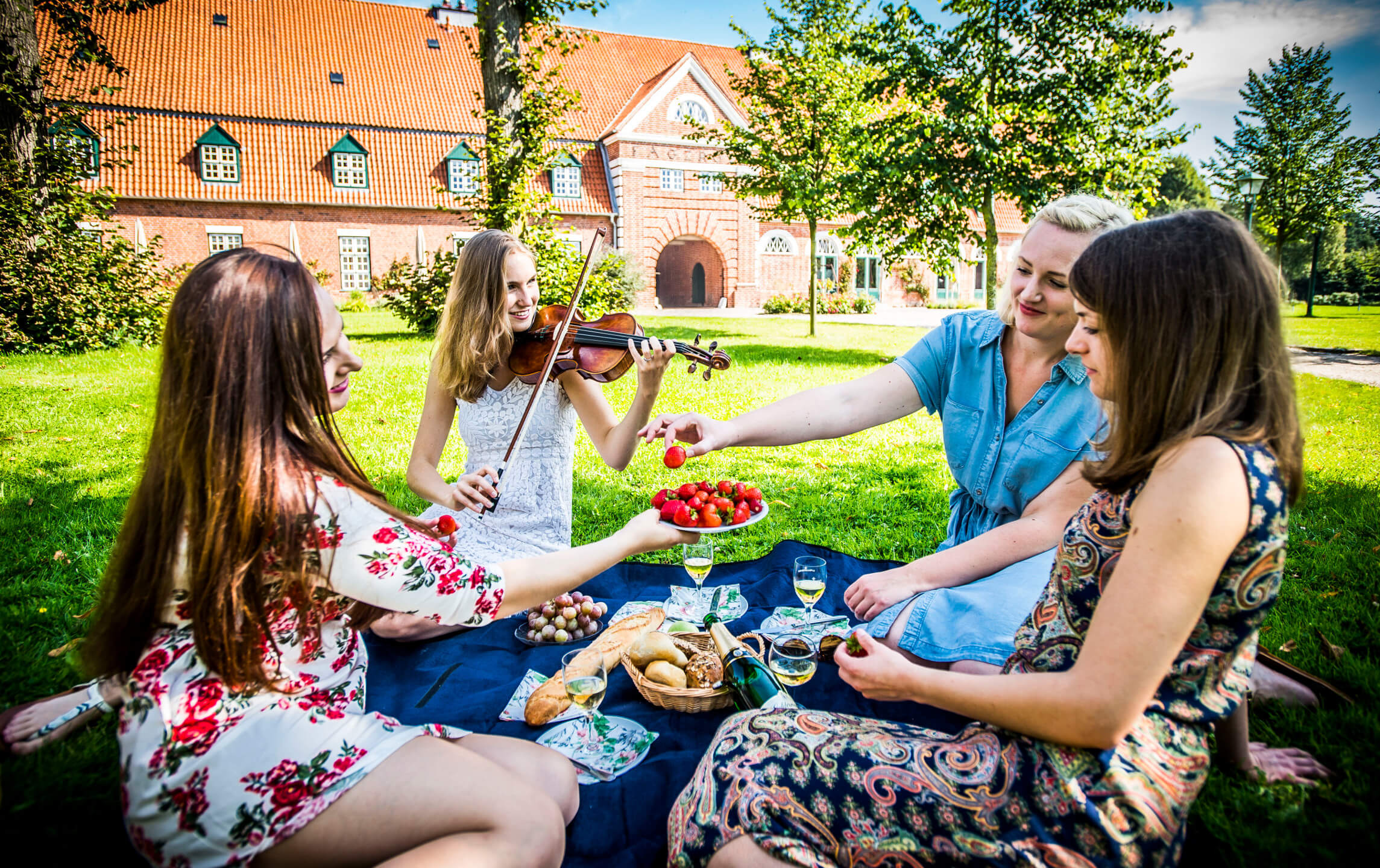Schleswig-Holstein Music Festival Four women are picnicking on a meadow in front of a brick building, one is playing the violin.