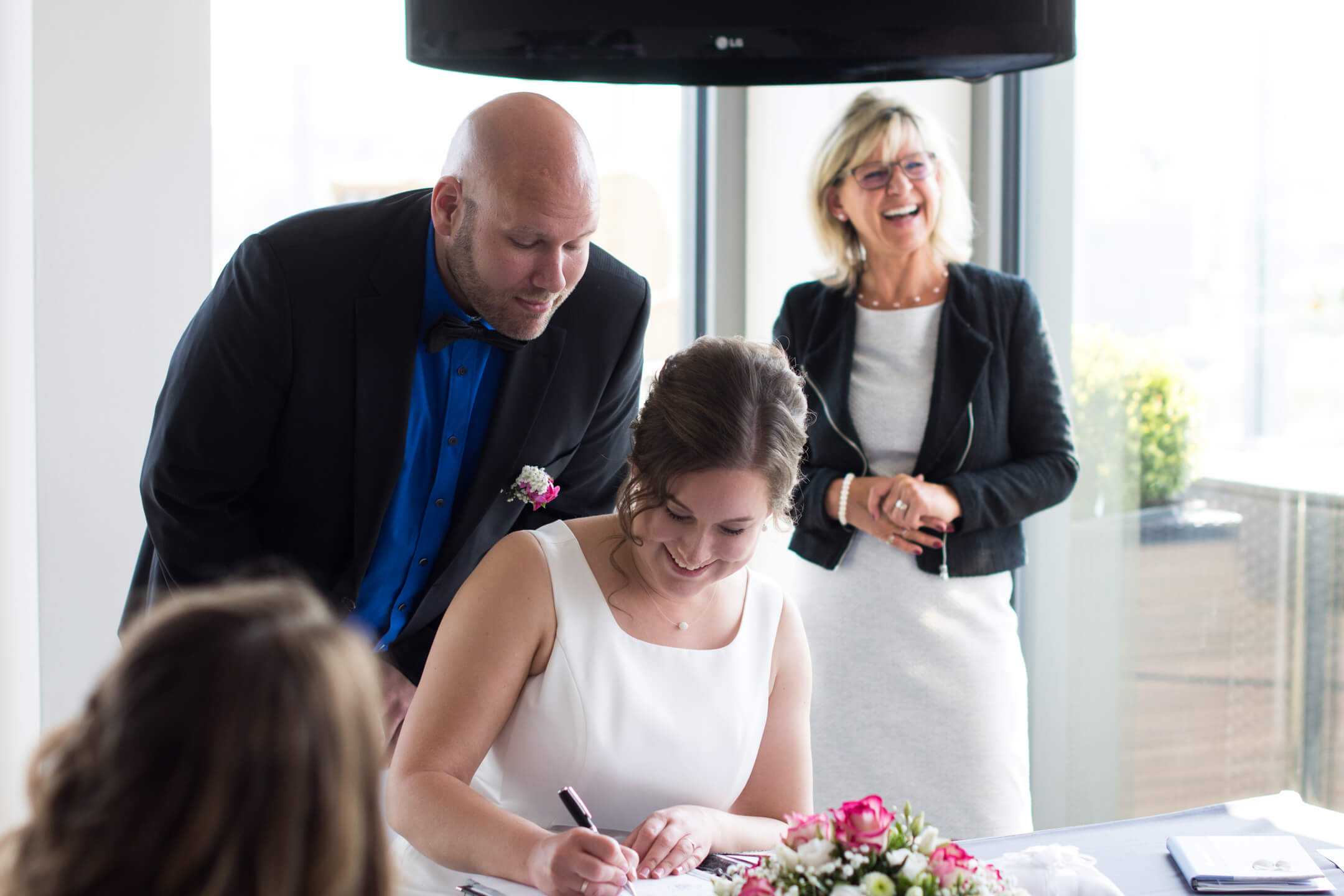 Brautpaar unterzeichnet Dokumente, während eine lächelnde Frau im Hintergrund steht, im ATLANTIC Hotel Kiel.