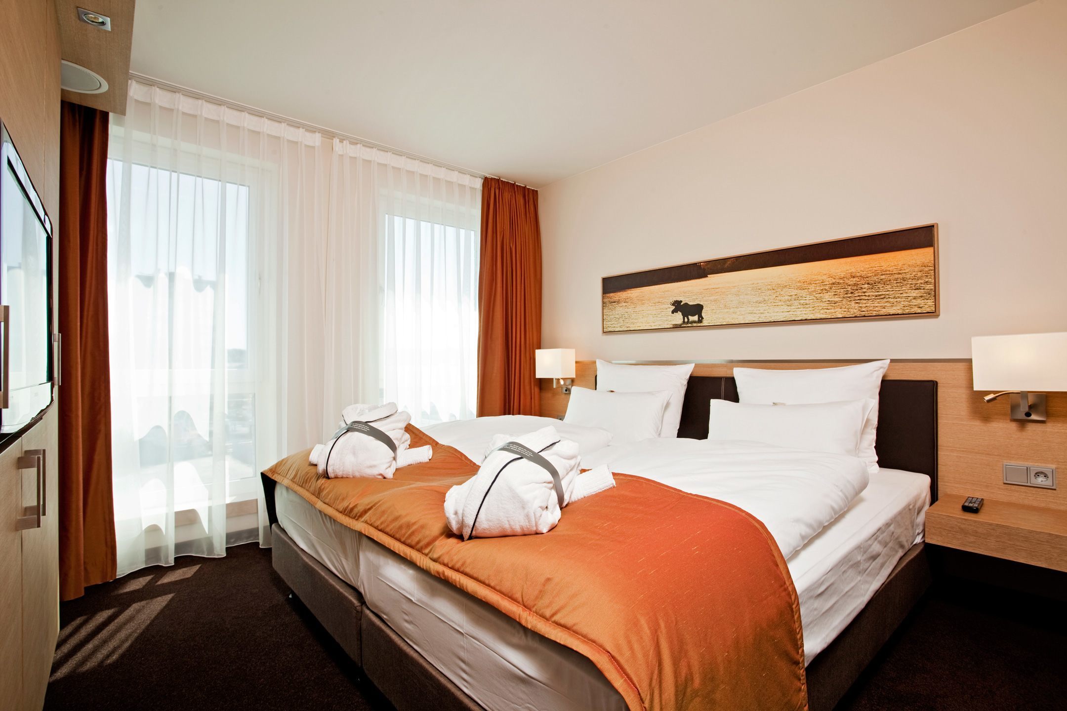 Modern hotel room with double bed, white sheets, orange blanket and bathrobes, light-colored curtains.