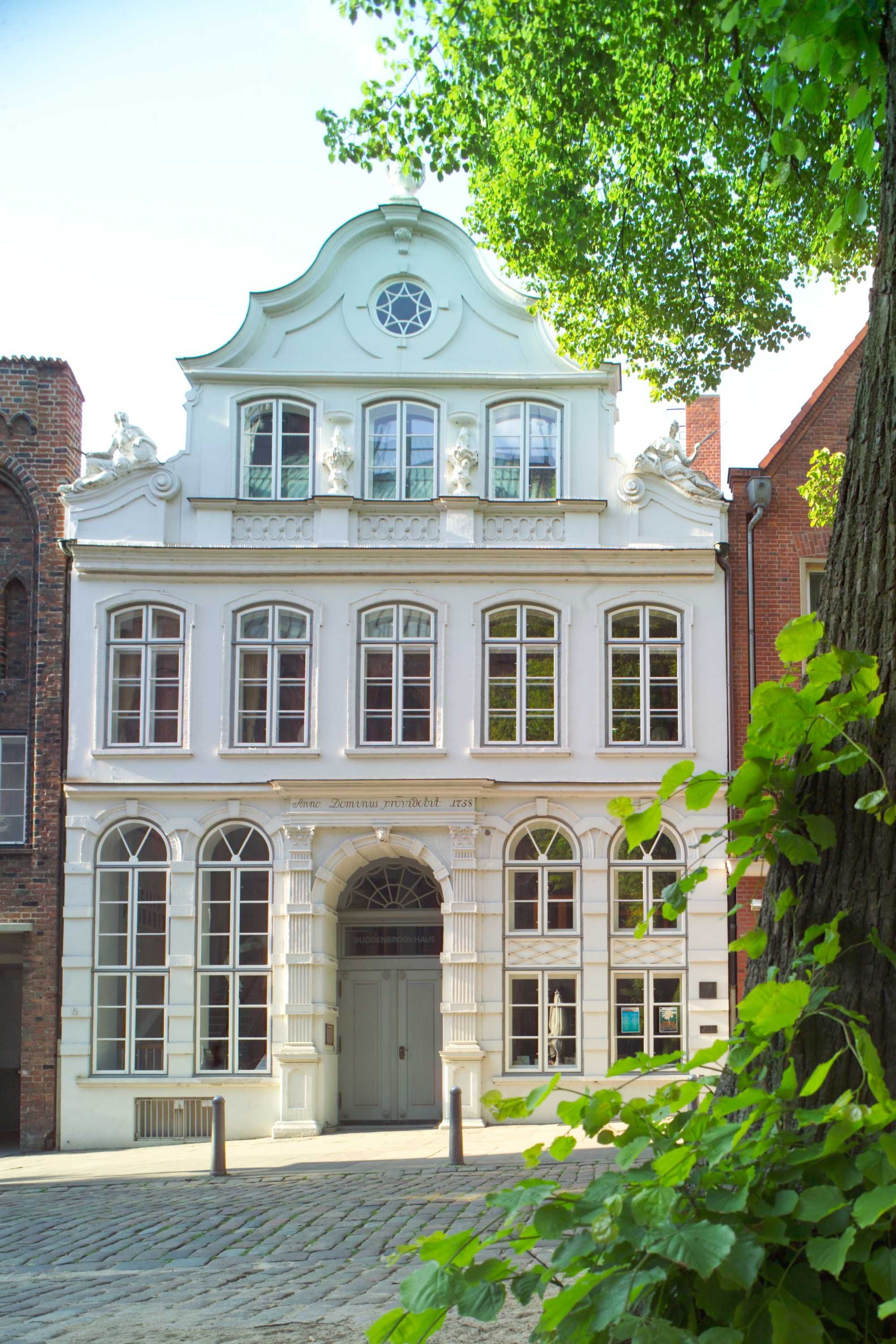 Historic, white façade of the Buddenbrookhaus in Lübeck, flanked by trees and cobblestones.