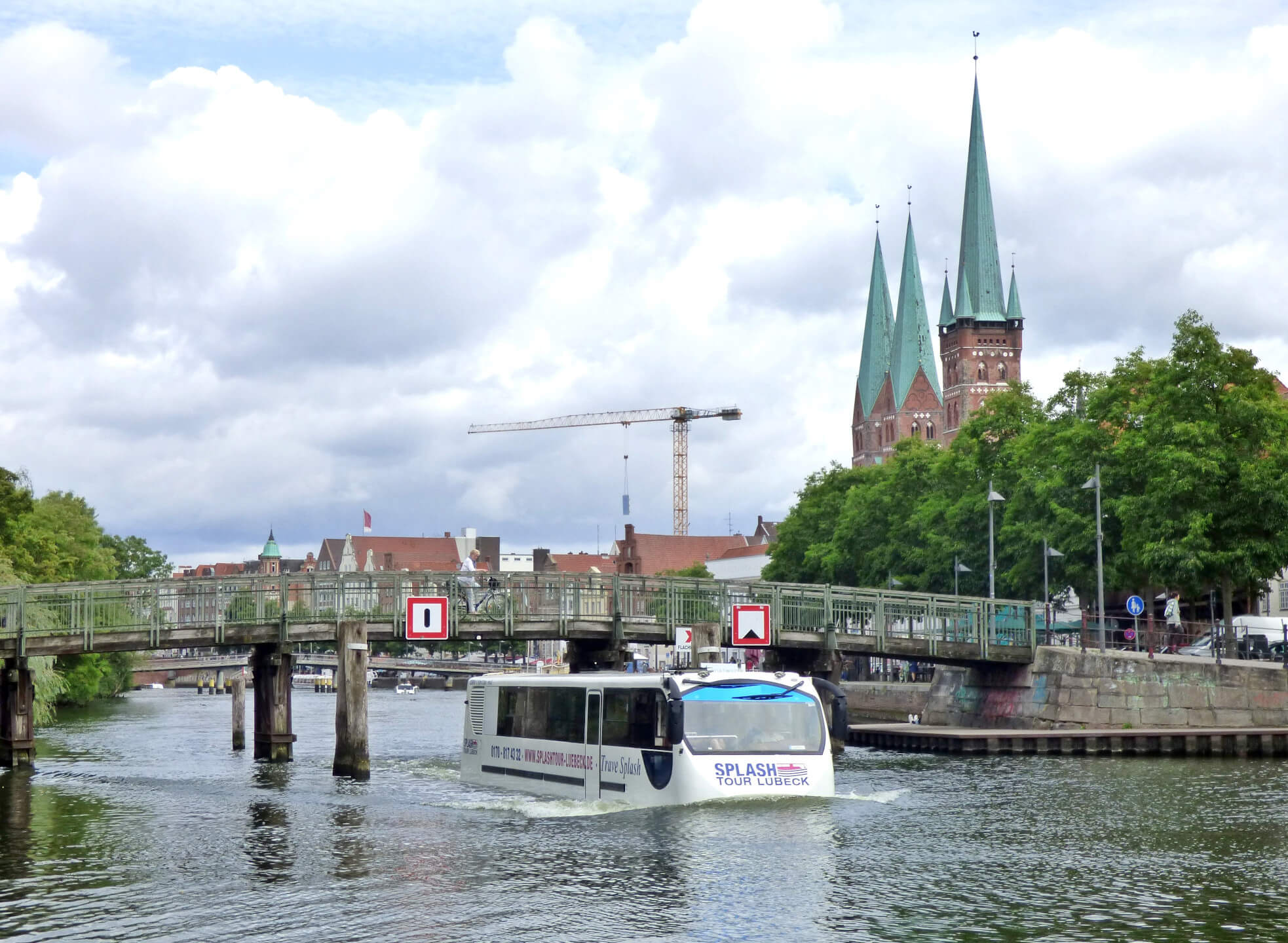 Boat on river in Lübeck, with bridge and church towers in the background, cloudy sky.