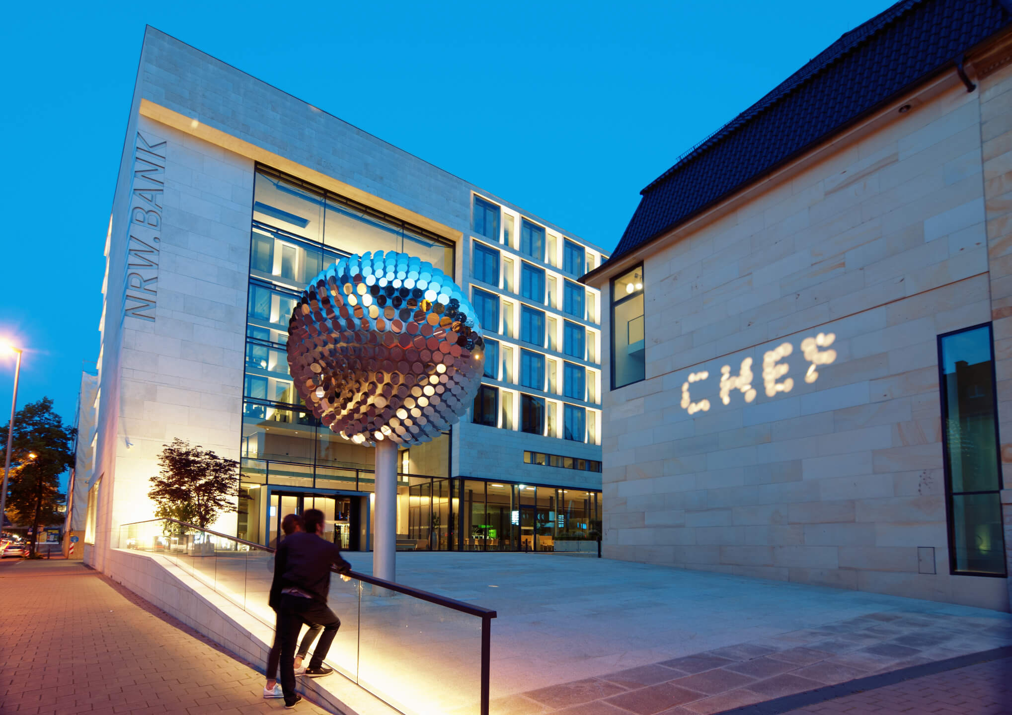 Sculpture Bit.Reflection by Julius Popp Modern NRW Bank building with illuminated façade and large, round sculpture at dusk.