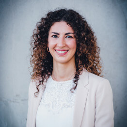 Eine lächelnde Frau mit lockigem Haar in einem hellen Blazer vor neutralem Hintergrund.