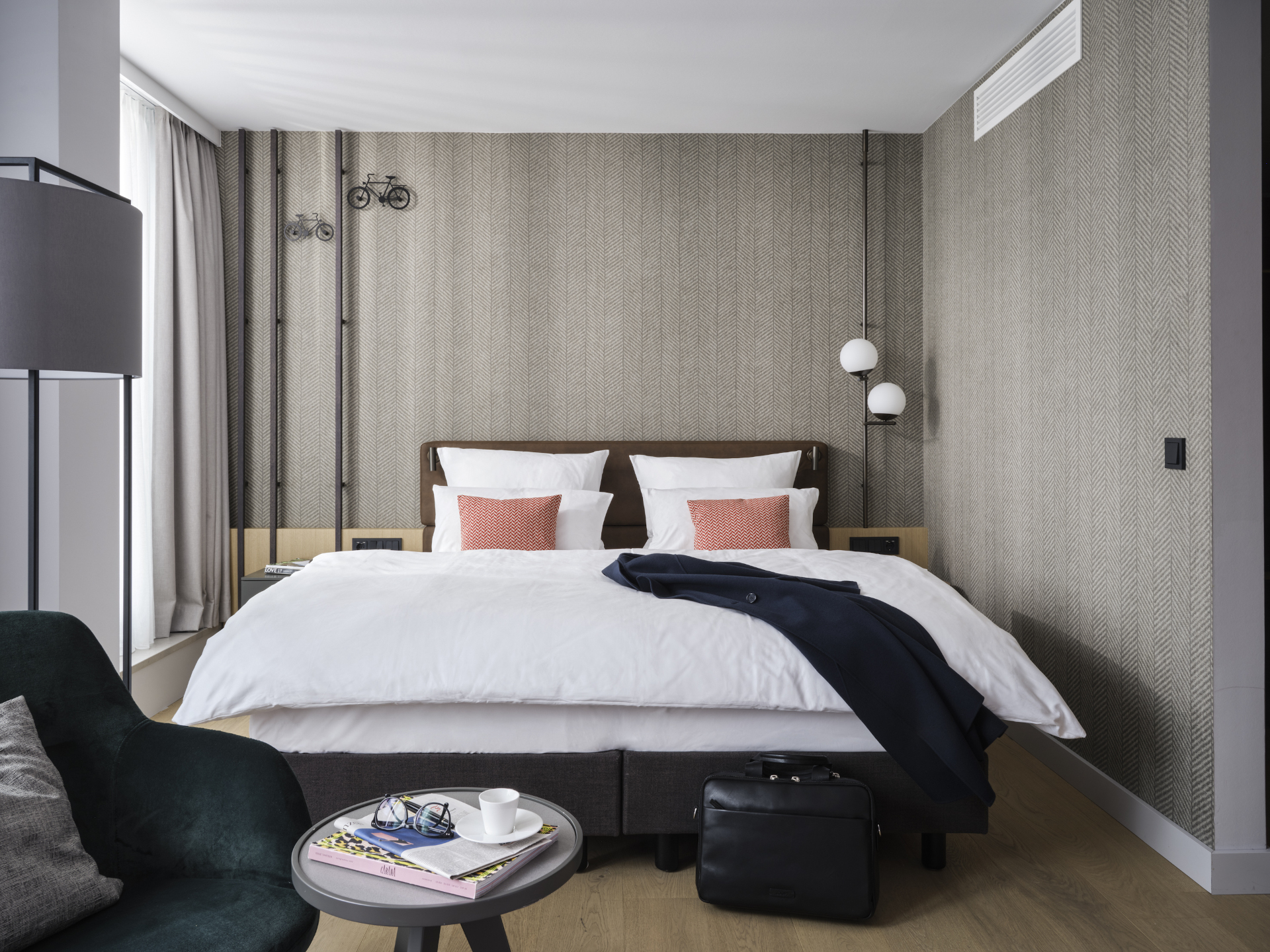 Modern hotel room with double bed, stylish lighting, armchair and table with glasses, cup and magazine.