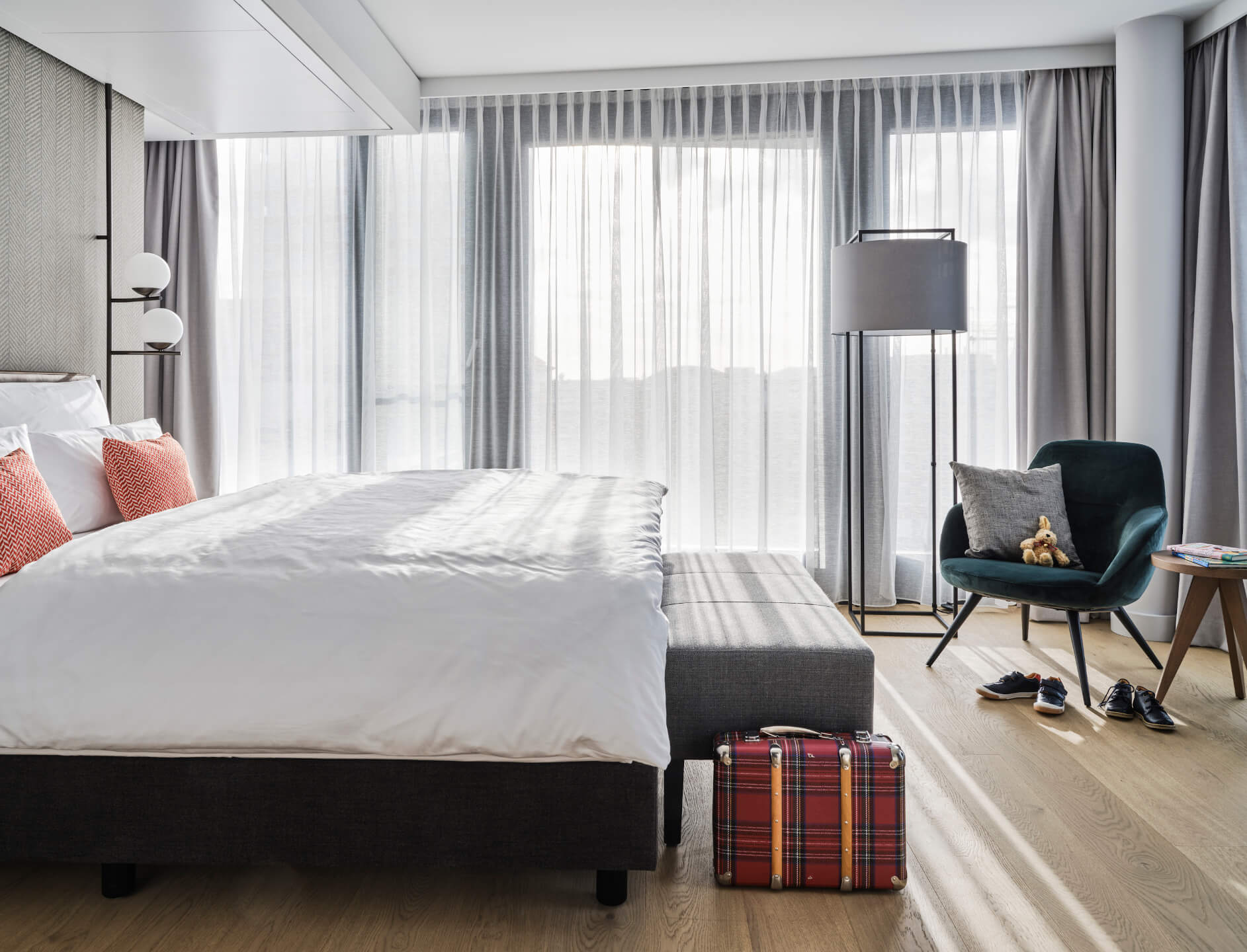 Helles Hotelzimmer mit großem Bett, roten Kissen, Sessel, Stehlampe und Koffer auf Holzfußboden.