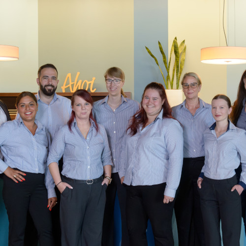 picture of the team from ATLANTIC Hotel Sail City Hotel staff at the ATLANTIC Hotel Sail City in Bremerhaven smile in uniforms in front of a sign reading "Ahoy".