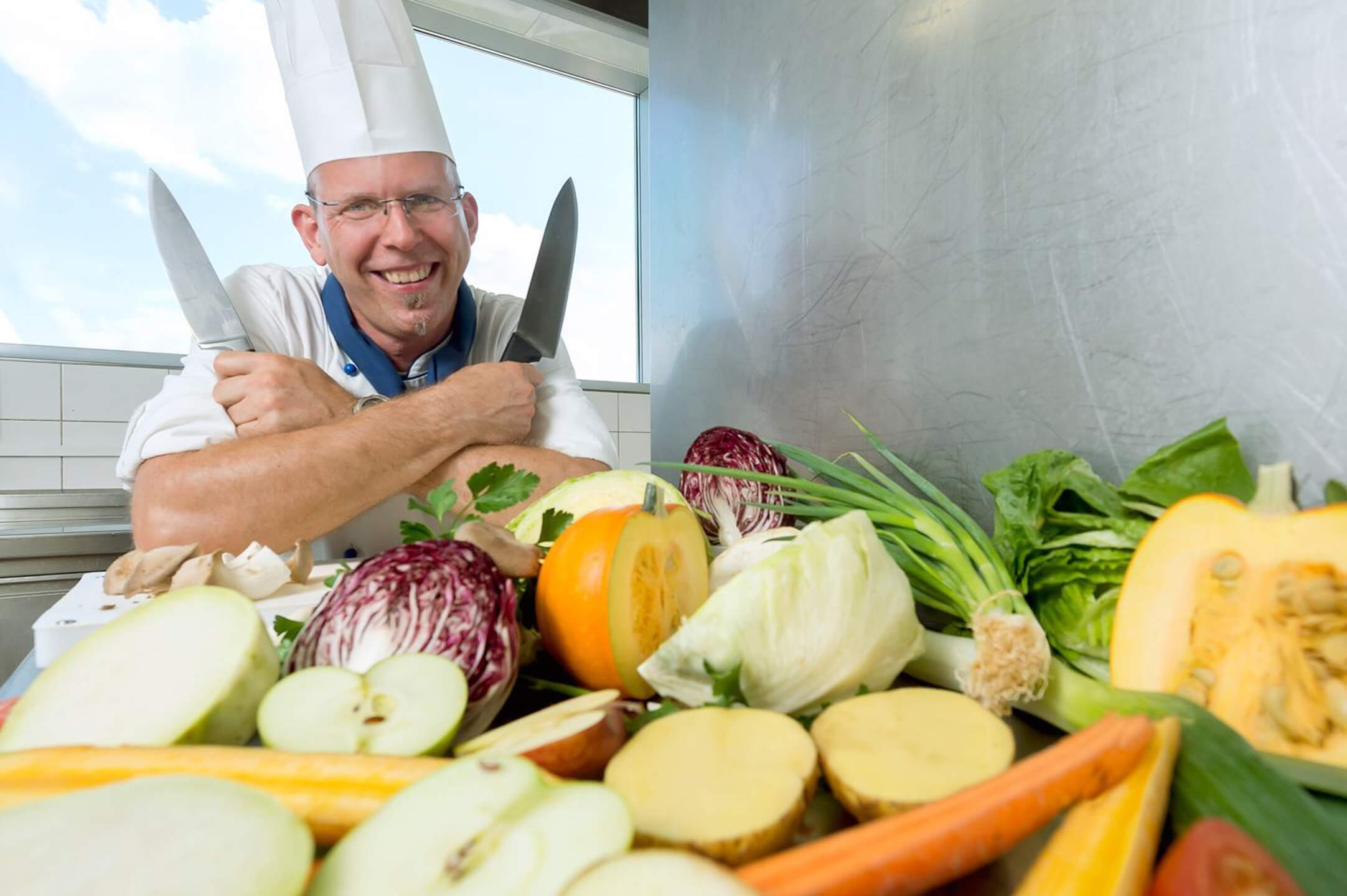 Atlantic Hotel Sail City – Green Sail Chef A smiling chef in uniform holds two knives surrounded by fresh vegetables in a hotel kitchen.