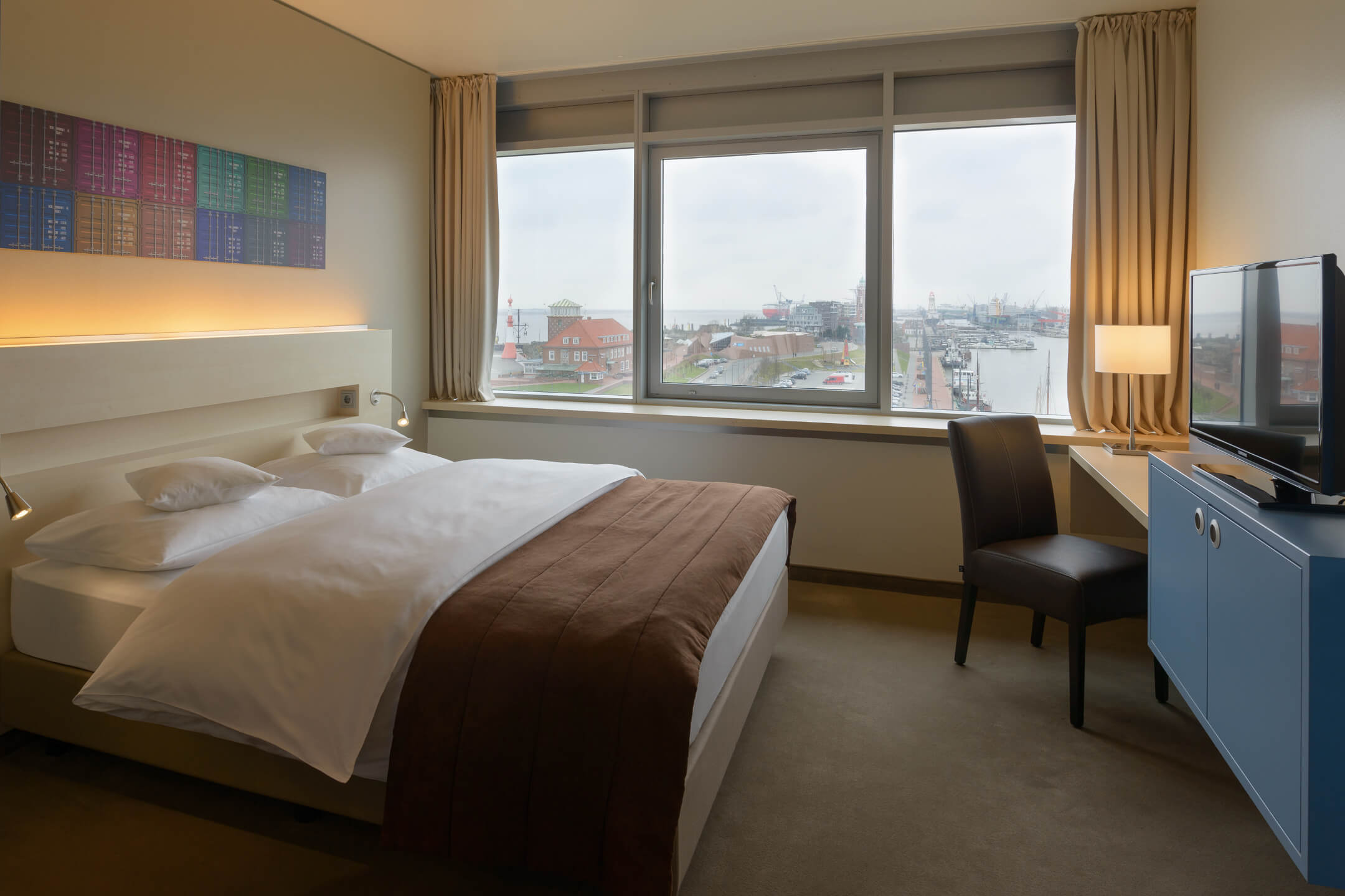 Bright hotel room with double bed, view of the harbor through large windows, TV and desk with lamp.