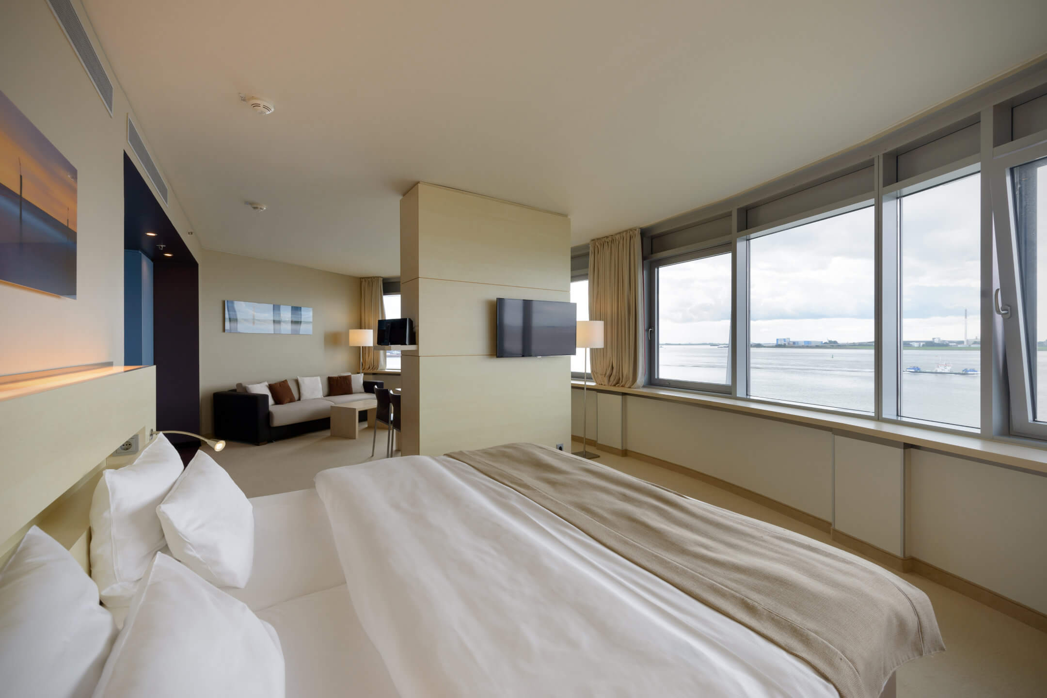 Helles Hotelzimmer mit großem Bett, Sofa, Fernseher und Panoramafenstern mit Blick auf das Wasser.