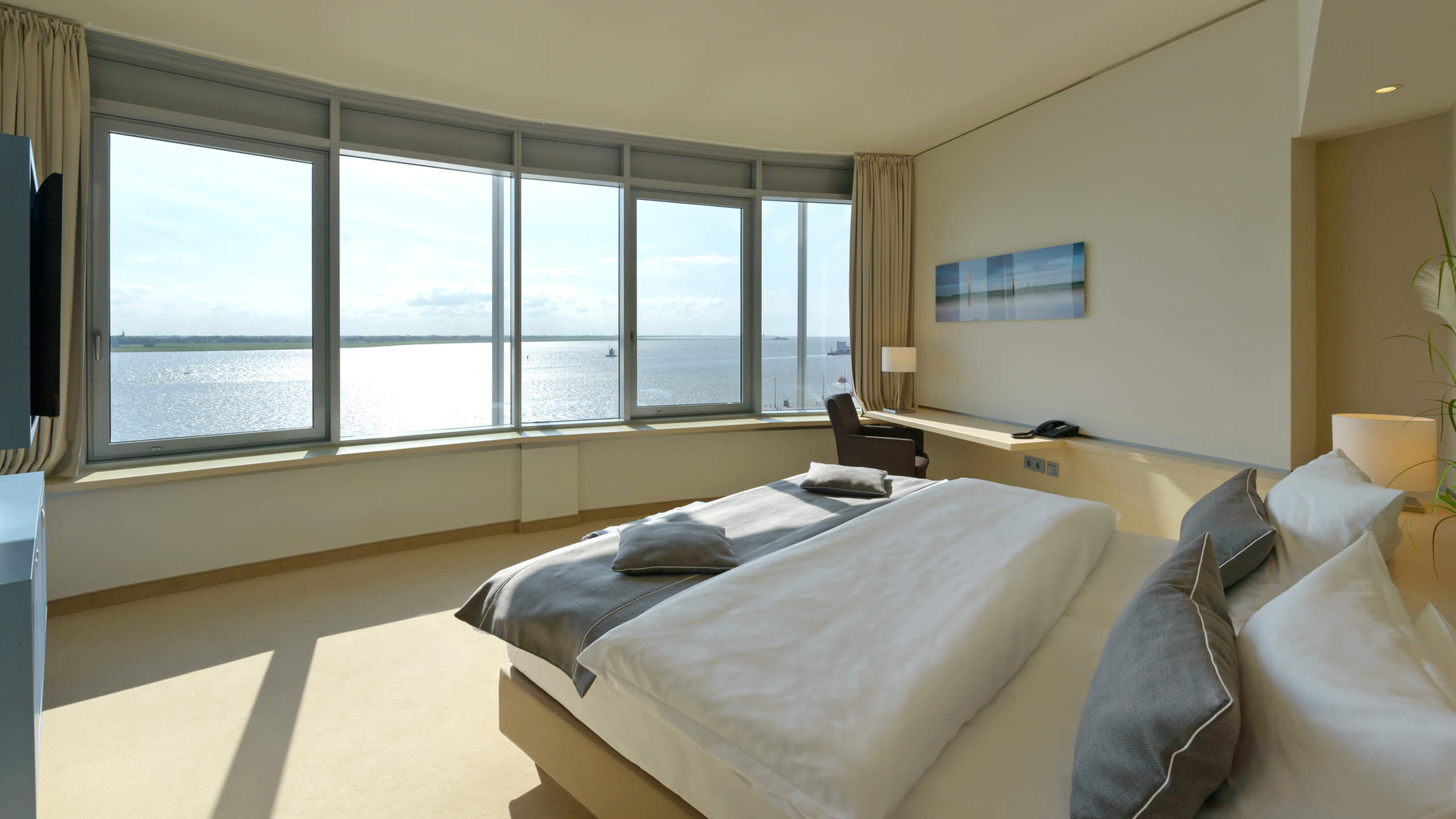 Bright hotel room with large bed and panoramic window overlooking the water.