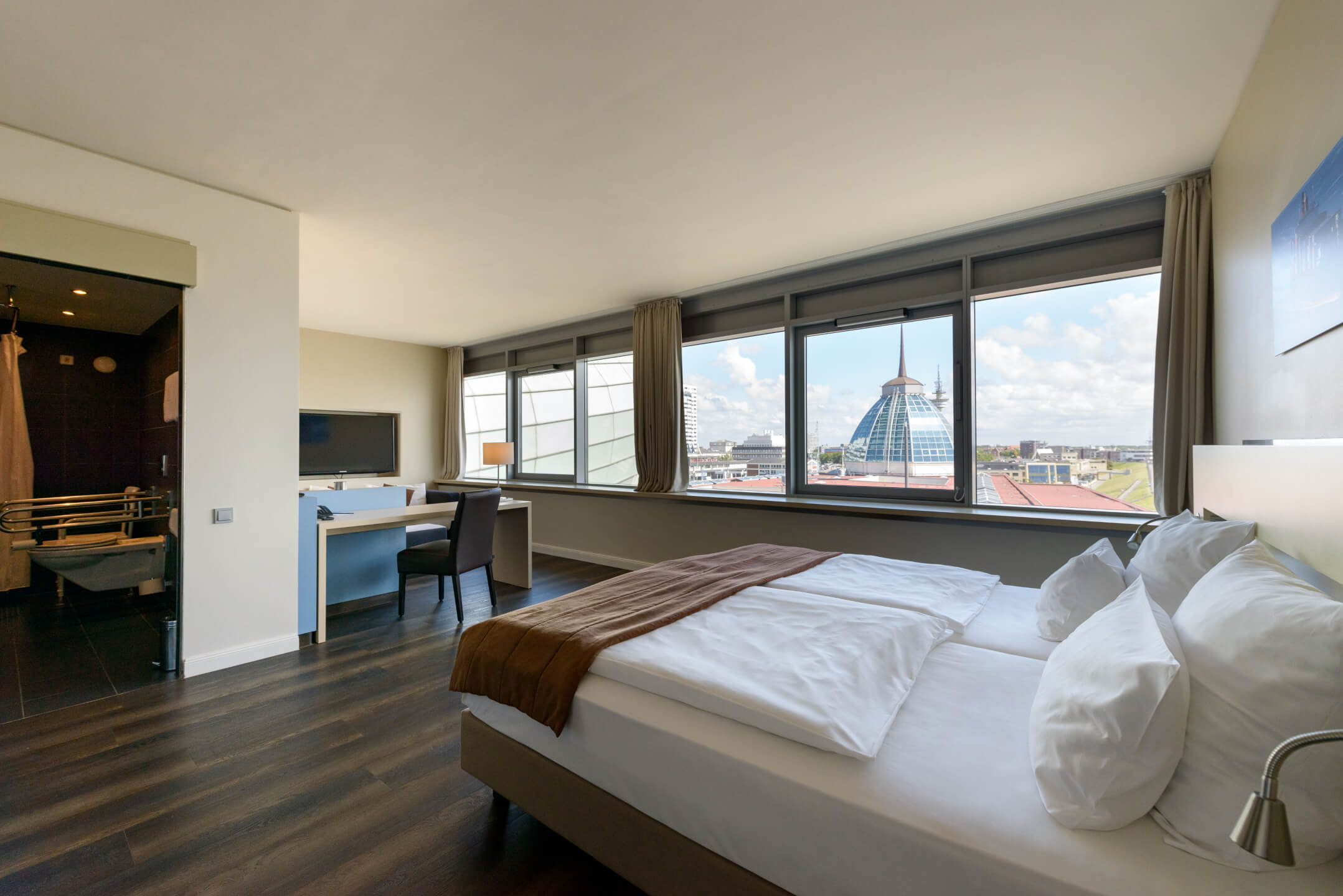 Modernes Hotelzimmer mit großem Fenster, Doppelbett, Schreibtisch und Blick auf die Stadtsilhouette von Bremerhaven.